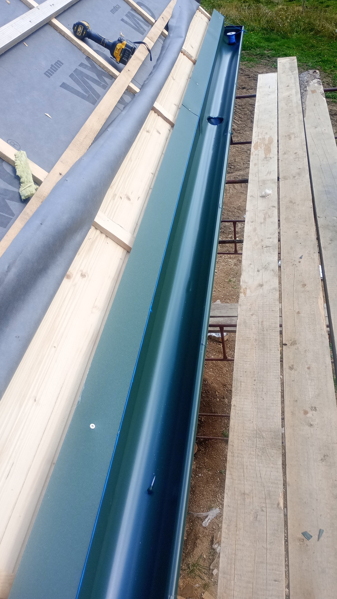 Montaż rynny dachowej w kolorze grafitowym, widok z góry. Na dachu widoczna membrana, drewniane łaty i wkrętarka. Rynna zamontowana, gotowa do odprowadzania wody.