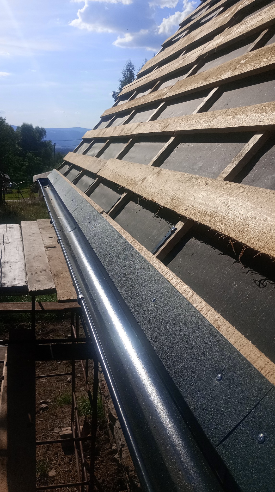 Montaż rynny w kolorze grafitowym na dachu z łatami i membraną, widok z dołu na tle błękitnego nieba i zielonych drzew. Detal mocowania rynny.