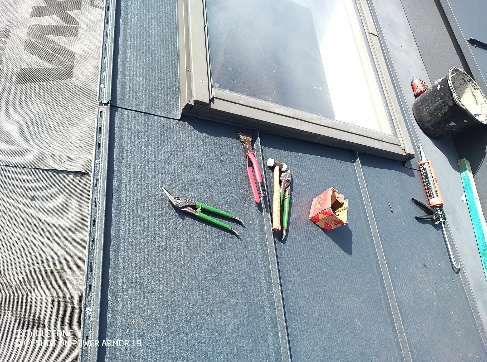 Szara blacha trapezowa na dachu z oknem. Widoczne narzędzia: nożyce do blachy, młotek, pędzel, pistolet do silikonu. Elementy montażowe i uszczelniające. Detal obróbki blacharskiej.