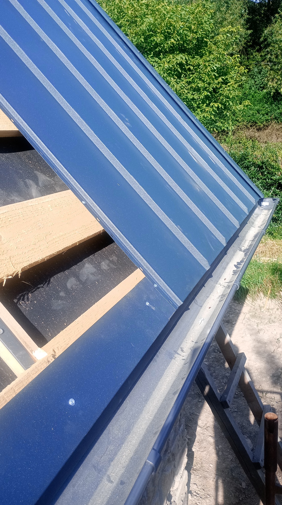 Niebieski dach z blachy trapezowej w trakcie montażu, widoczne drewniane elementy konstrukcji i zamontowana rynna. Ujęcie z dołu, na tle zieleni.