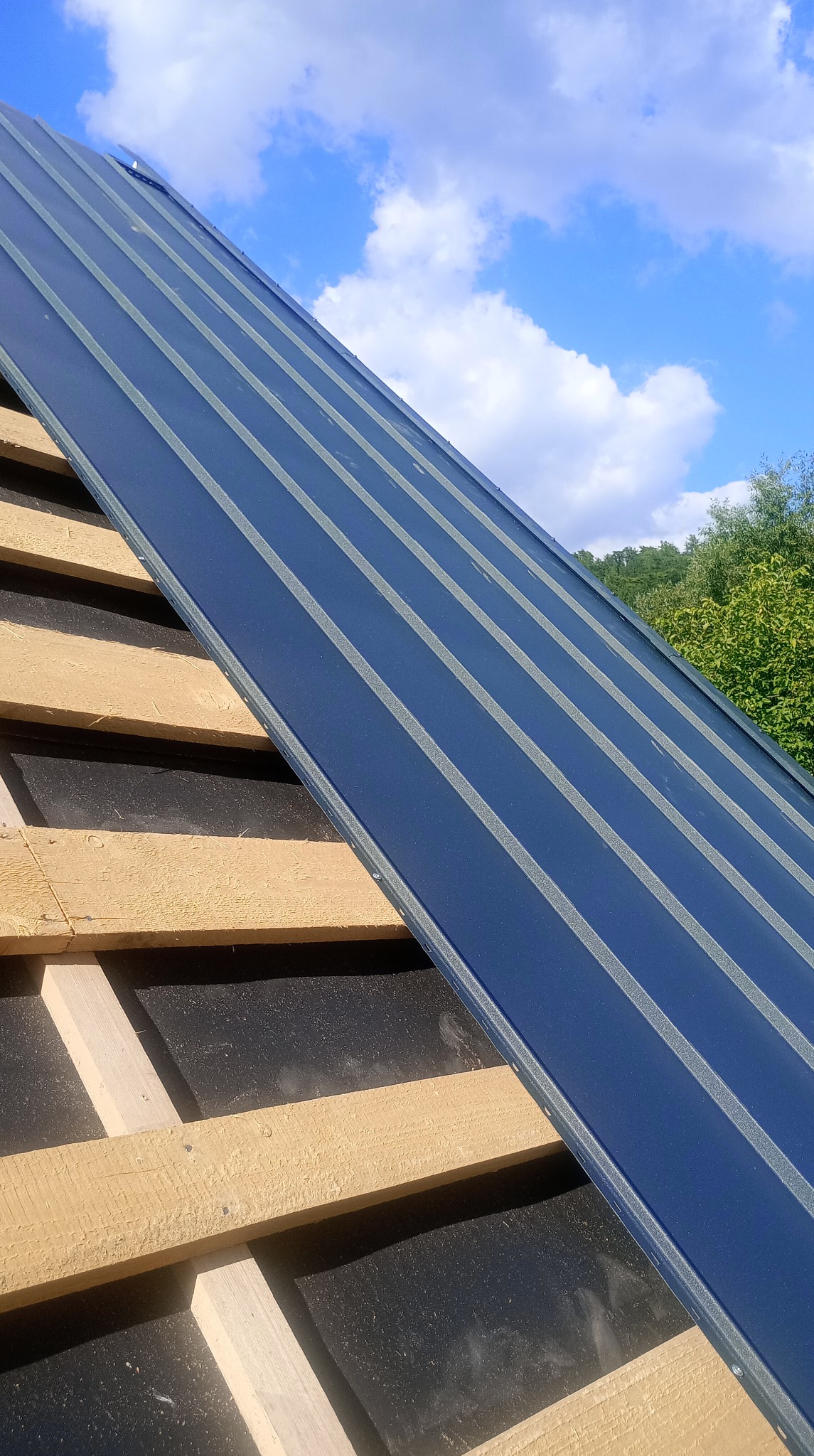 Niebieskie panele dachowe układane na drewnianej konstrukcji dachu na tle błękitnego nieba z chmurami i zieleni drzew. Widok z dołu, ukośny kadr.