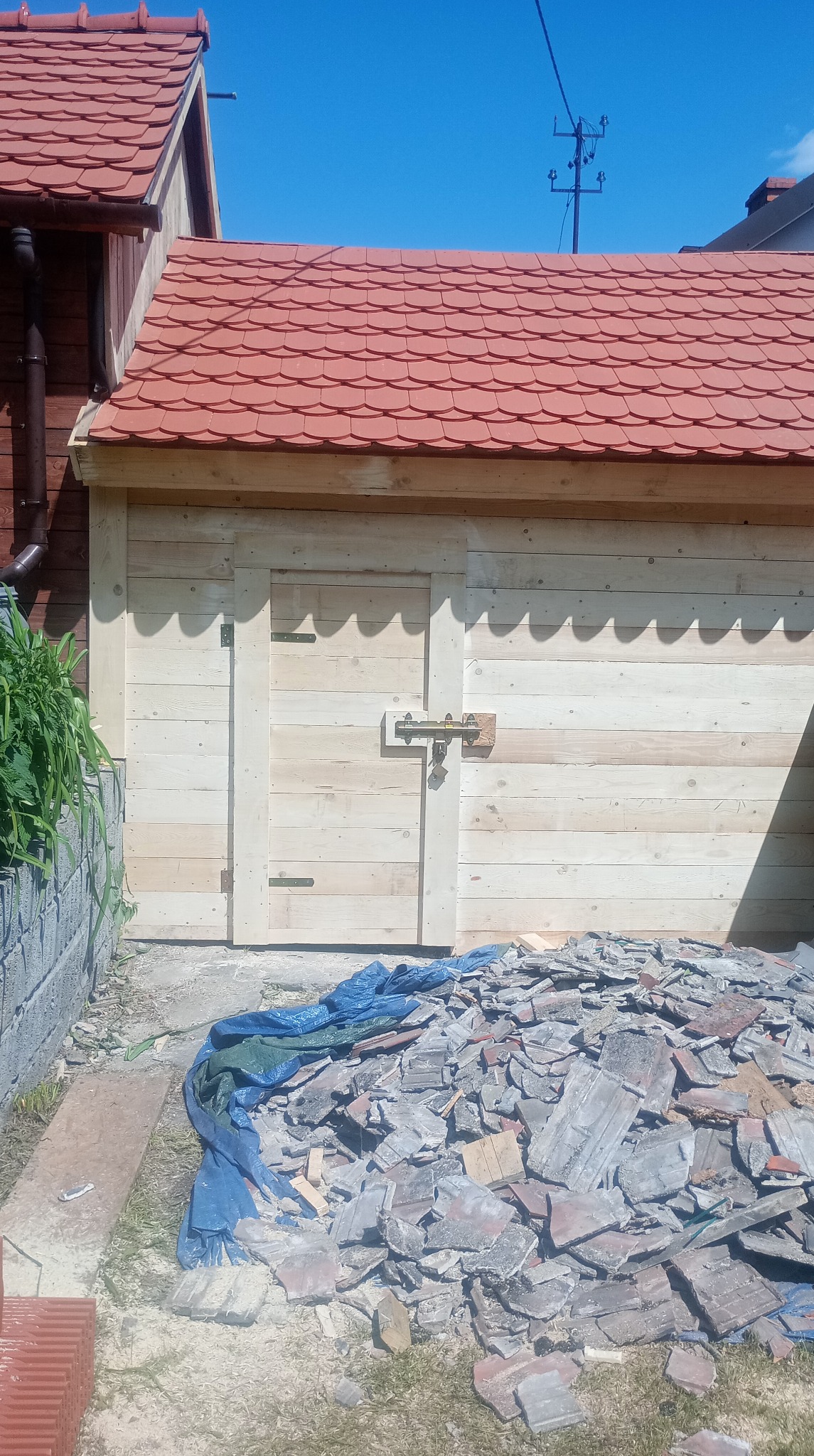 Nowy, czerwony dach z dachówki karpiówki na drewnianym budynku gospodarczym. Sterta starych dachówek na ziemi. Widok na błękitne niebo i słupy energetyczne.