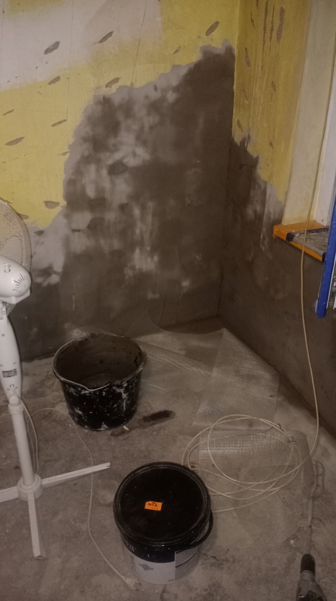 Fragment ściany po nałożeniu szarej zaprawy, widoczne siatki wzmacniające, dwa wiadra z zaprawą i wentylator. Etap prac przygotowawczych do wykończenia ścian.