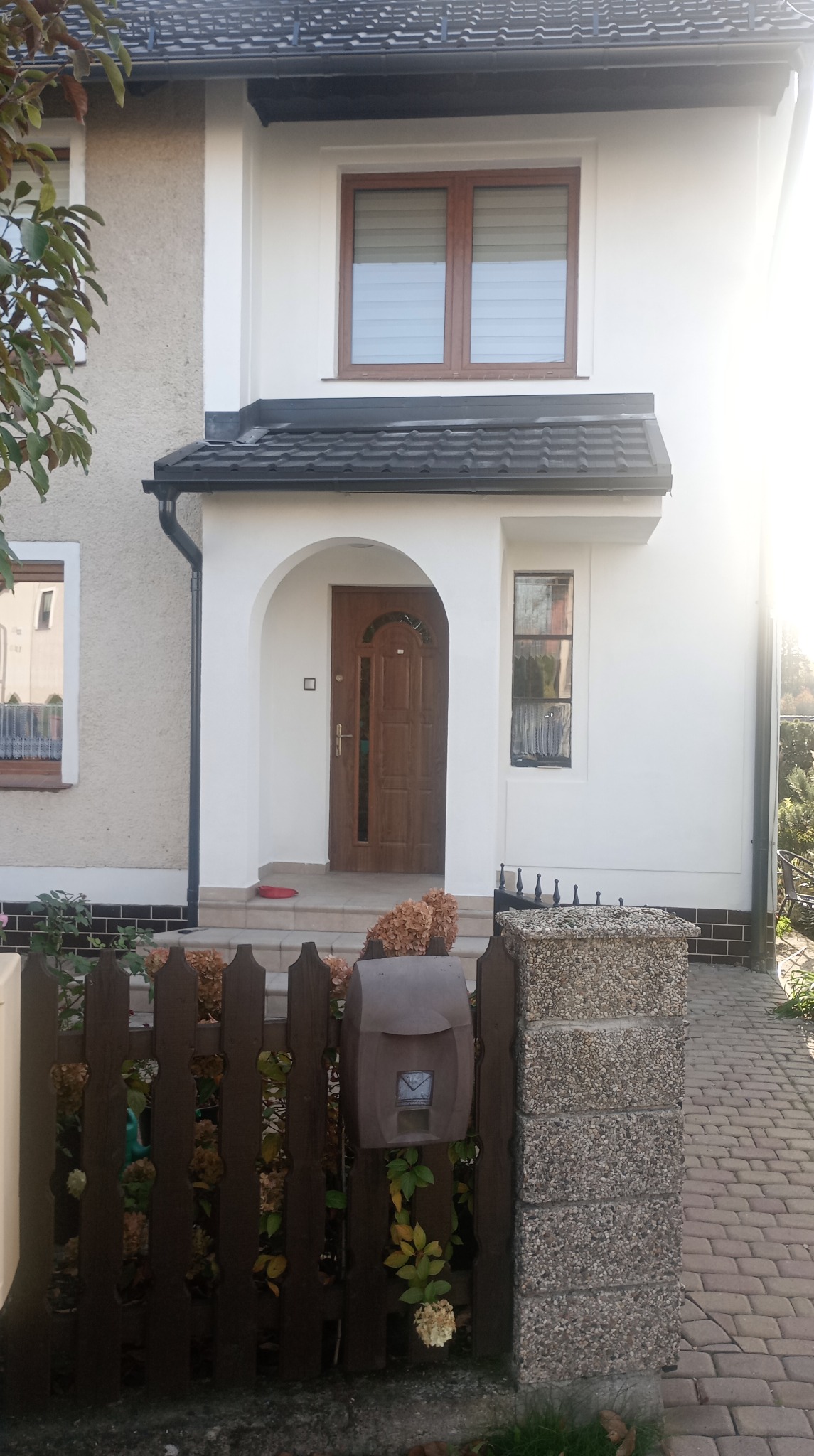 Dwukondygnacyjny dom z ciemną dachówką i rynnami, kremową elewacją i brązową stolarką okienną. Widoczny fragment ogrodzenia z skrzynką na listy.