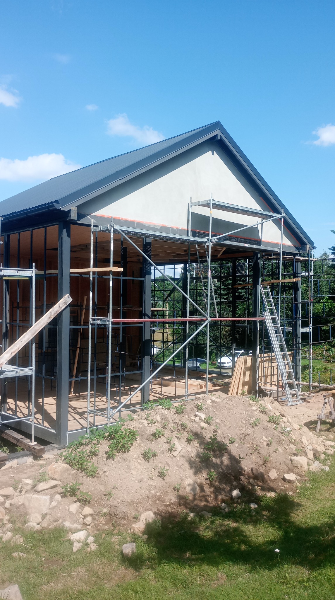 Budowa domu w toku: widok dachu z szarej blachy trapezowej i rusztowań na tle błękitnego nieba. Konstrukcja szkieletowa z czarnych profili.