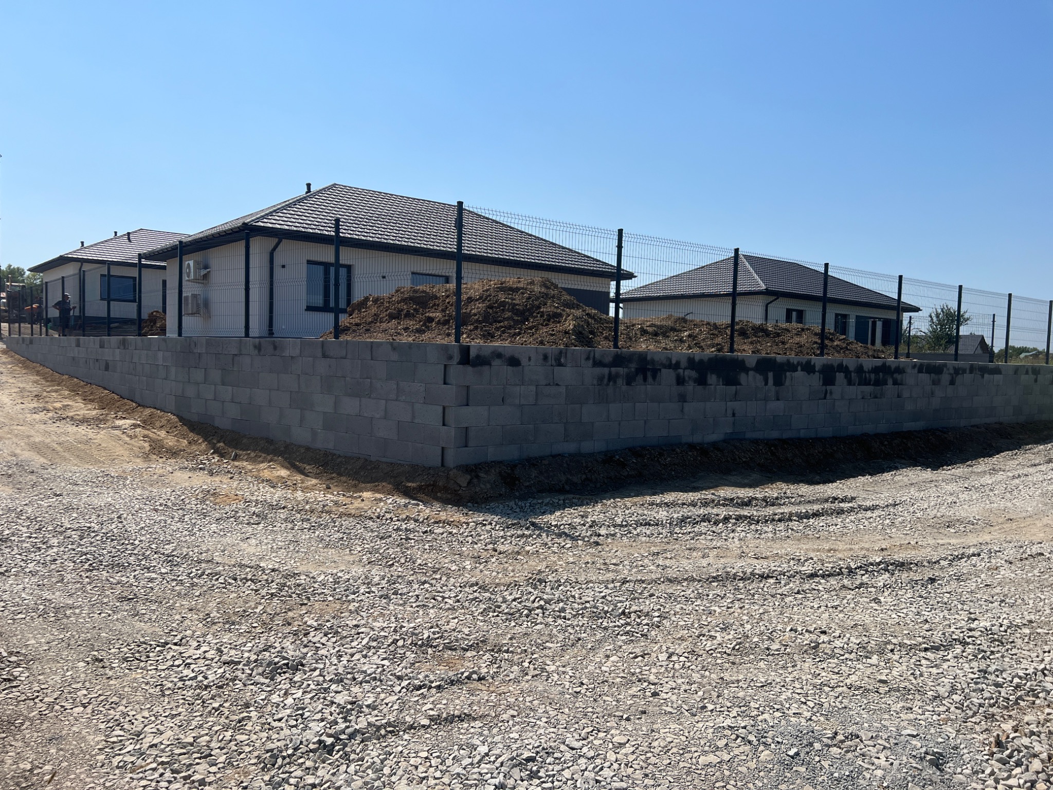 Nowoczesne domy jednorodzinne z szarymi dachami, ogrodzone panelami na podmurówce z bloczków betonowych. Teren wokół w trakcie prac ziemnych, widoczny gruz.