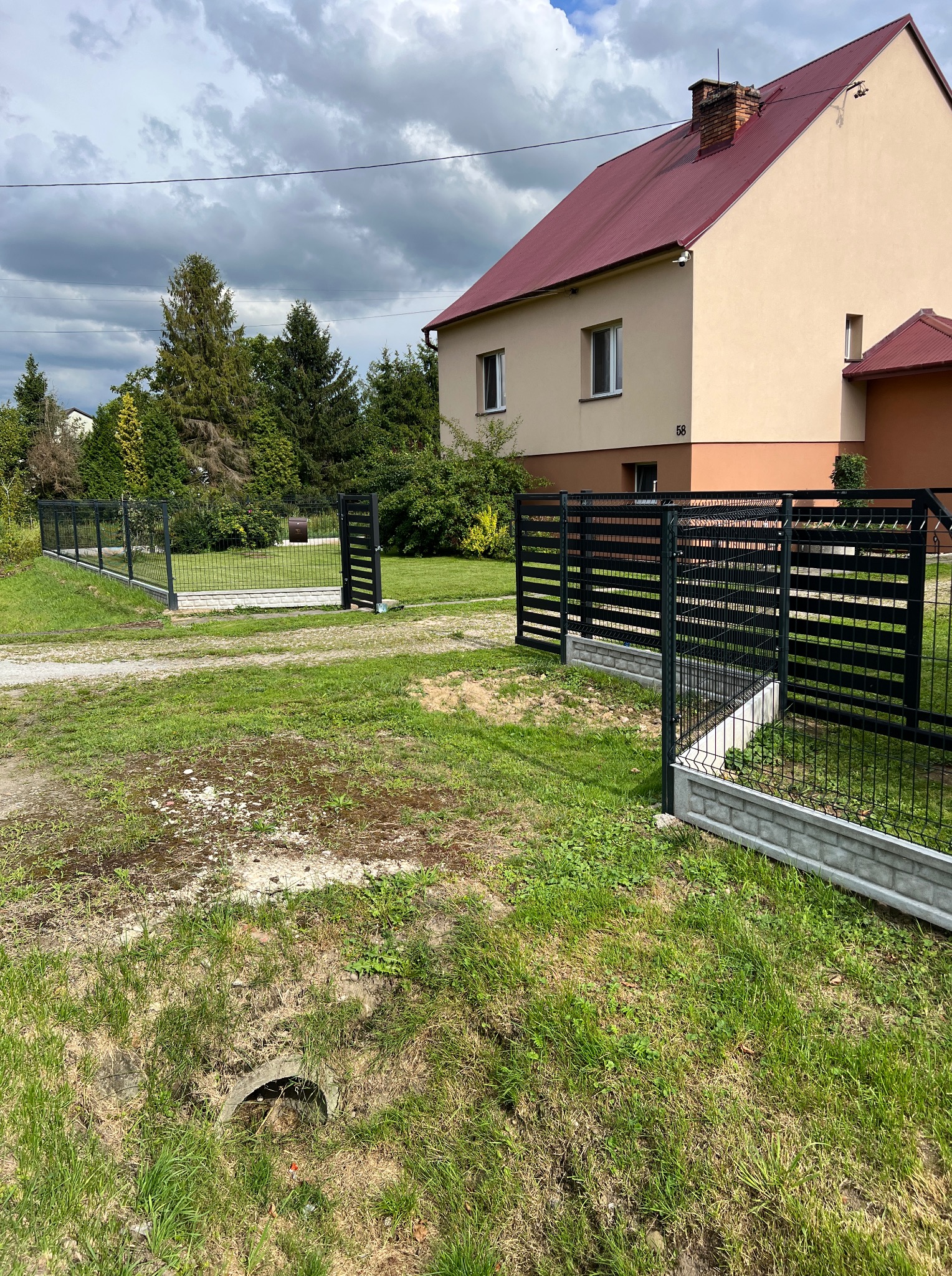 Nowoczesne ogrodzenie panelowe z bramką na tle domu z czerwonym dachem. Elementy poziome w bramce i ogrodzeniu. Widok na posesję z zewnątrz.