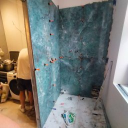 Marcin Wasiak - Montaż płytek imitujących marmur w kabinie prysznicowej z użyciem klipsów poziomujących. Widoczna drabina i częściowo osoba wykonująca prace.