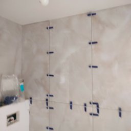 Marcin Wasiak - Ściana łazienki w trakcie układania płytek w odcieniach beżu, z widocznymi niebieskimi krzyżykami dystansowymi i sznurkiem poziomującym. Fragment instalacji podtynkowej.