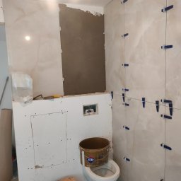 Marcin Wasiak - Wnętrze łazienki w trakcie prac glazurniczych: ułożone płytki imitujące marmur z niebieskimi krzyżykami dystansowymi, przygotowana ściana pod płytki.