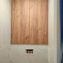 Wykończona toaleta podtynkowa z drewnianą szafką i marmurowymi płytkami. Widoczna instalacja w ścianie przed montażem przycisku spłukującego.