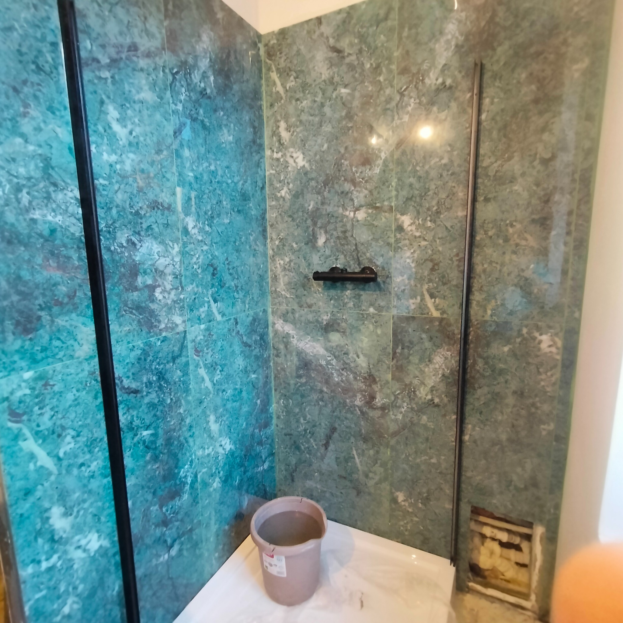 Wykończona kabina prysznicowa z zielonymi, marmurkowymi płytkami. Widoczne czarne ramy kabiny i akcesoria. Na podłodze wiadro i niedokończone miejsce na rurę.