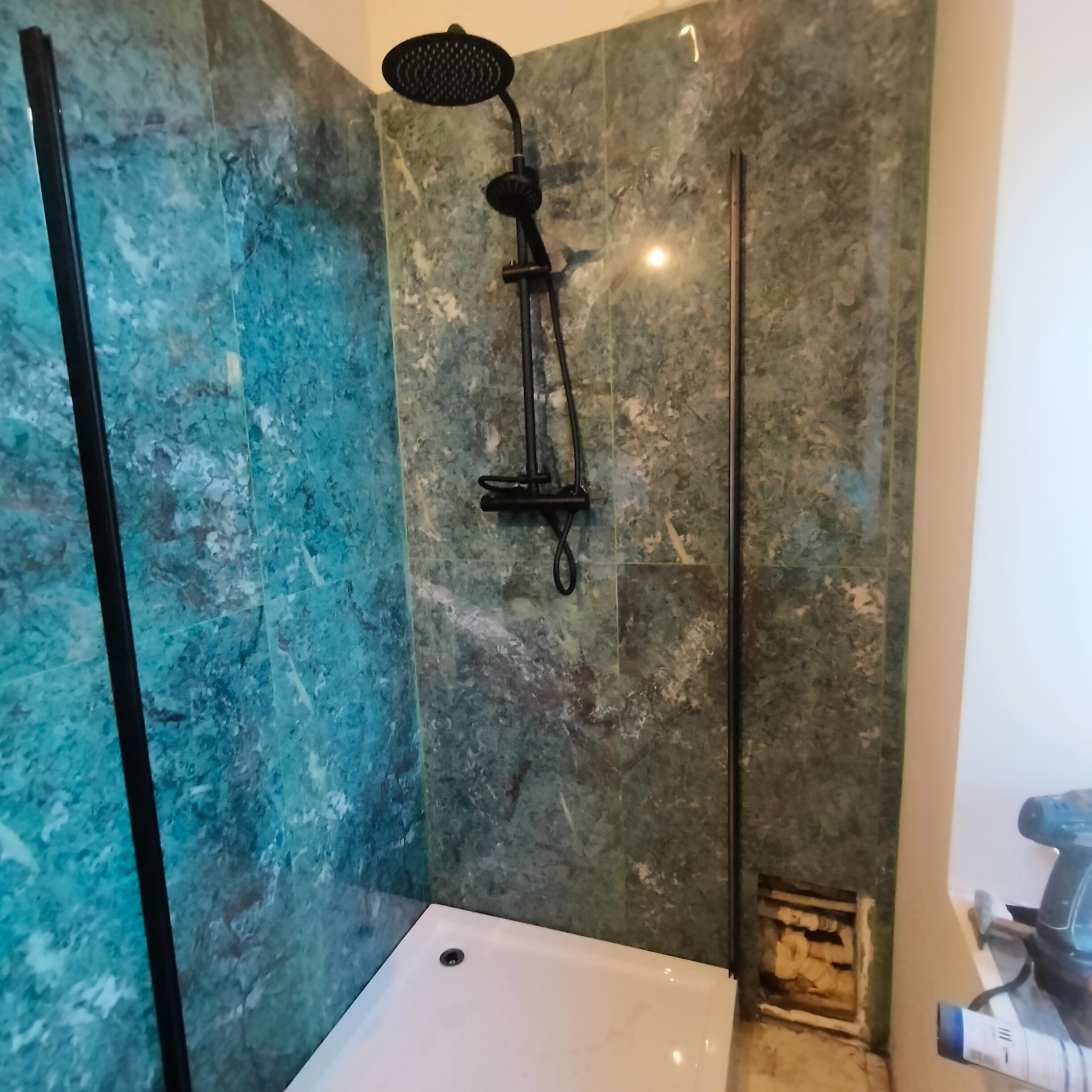Wykończona kabina prysznicowa z panelami imitującymi marmur w odcieniach zieleni i błękitu, czarna armatura, biały brodzik. Widoczny fragment instalacji.