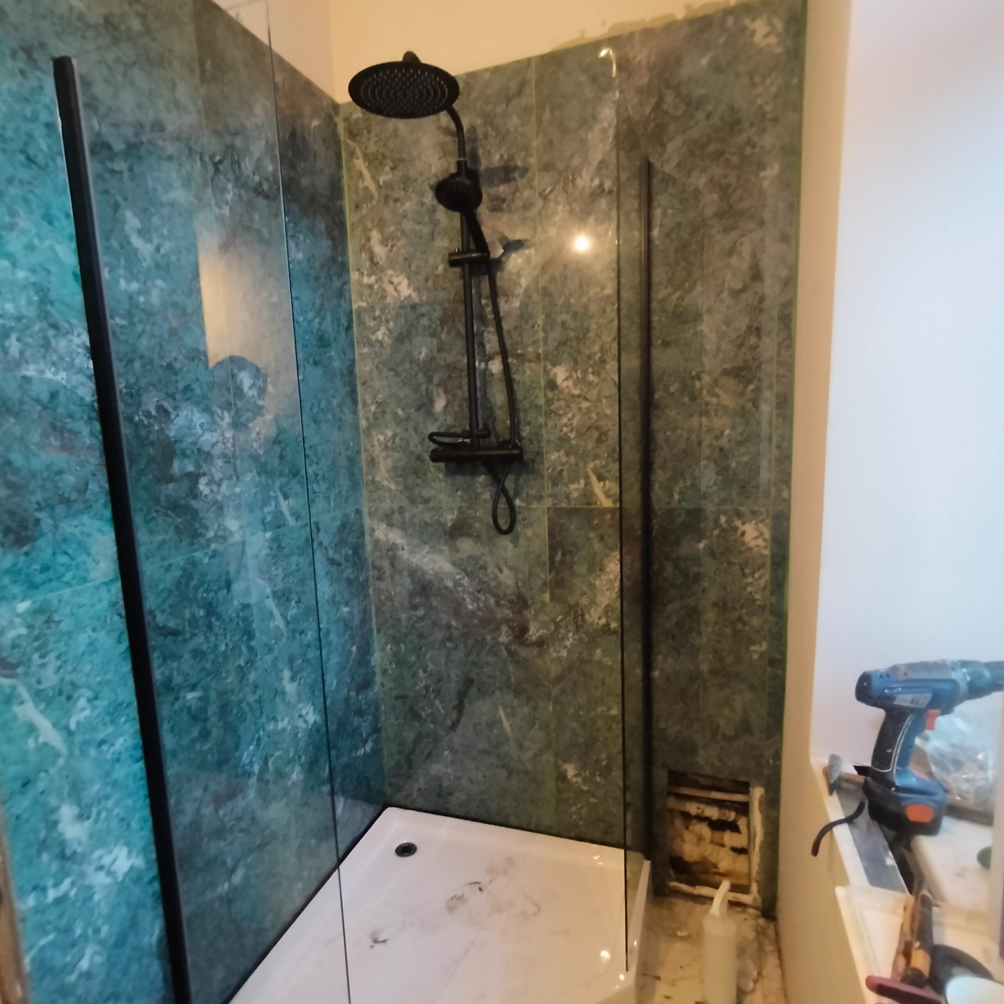 Kabina prysznicowa z deszczownicą i zielonymi płytkami imitującymi marmur. W tle widać narzędzia, co sugeruje trwający remont łazienki.
