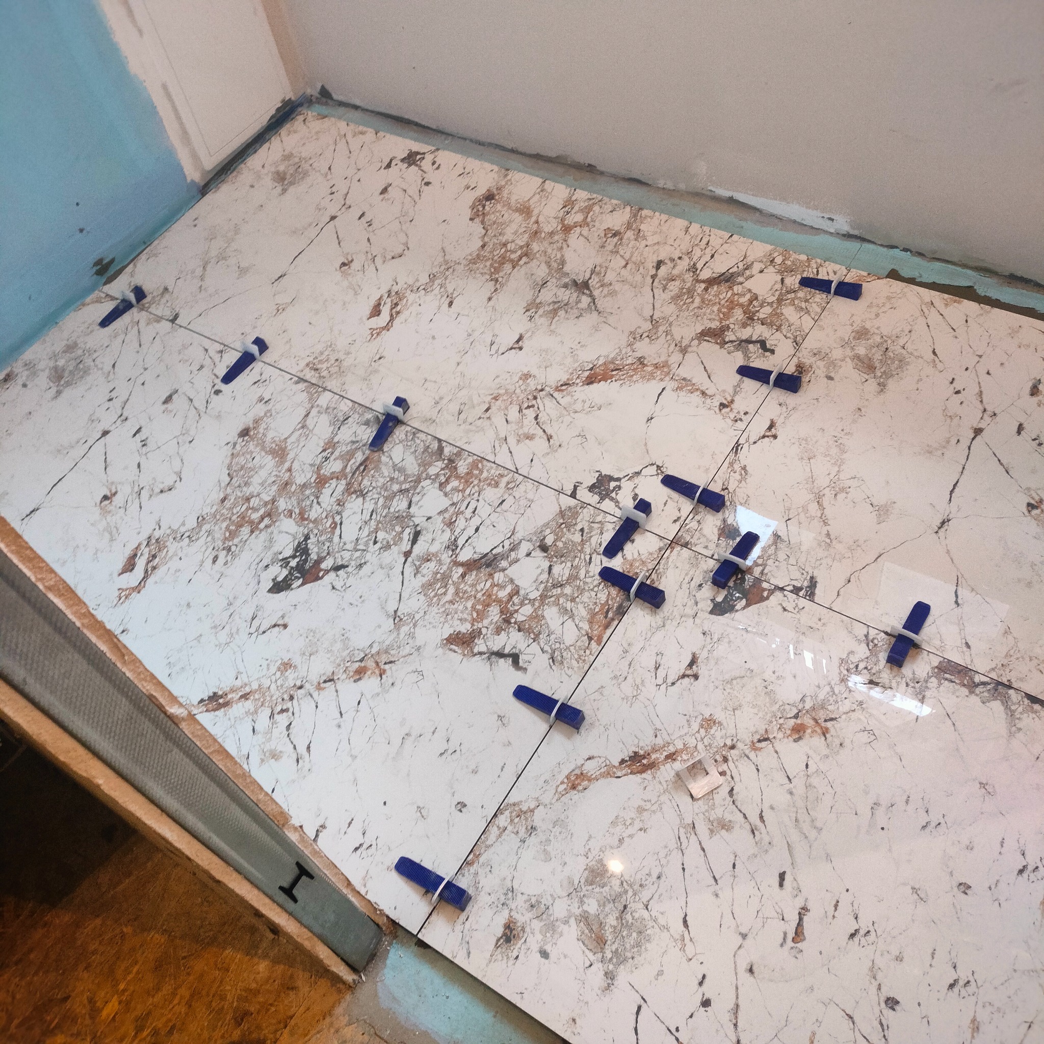 Układanie płytek imitujących marmur, z niebieskimi krzyżykami dystansowymi, na podłodze w trakcie remontu. Widoczne profile i fragment ściany.