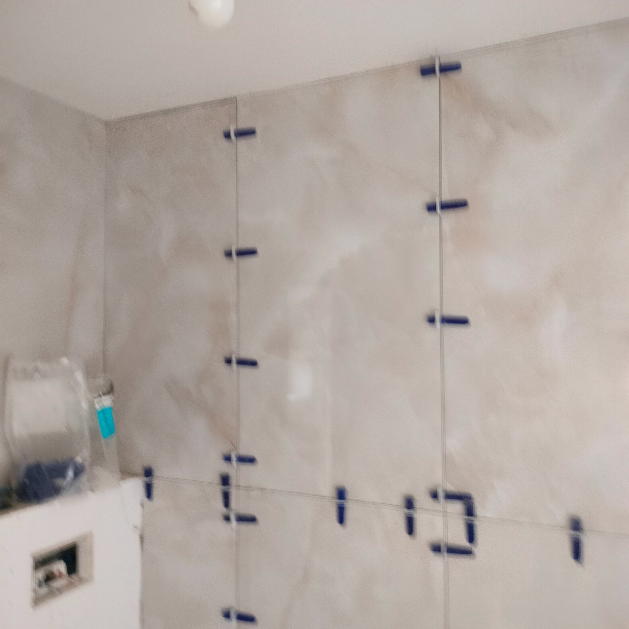 Ściana łazienki w trakcie układania płytek w odcieniach beżu, z widocznymi niebieskimi krzyżykami dystansowymi i sznurkiem poziomującym. Fragment instalacji podtynkowej.