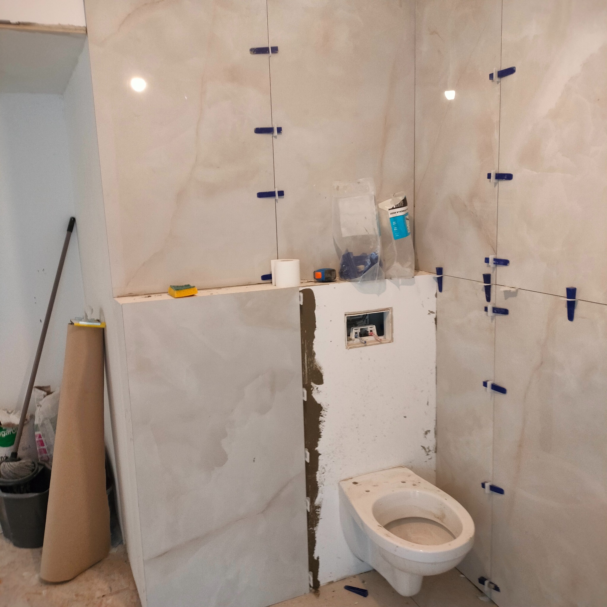 Łazienka w trakcie prac glazurniczych: beżowe płytki z niebieskimi krzyżykami dystansowymi, częściowo zamontowana toaleta, widoczne narzędzia i materiały budowlane.