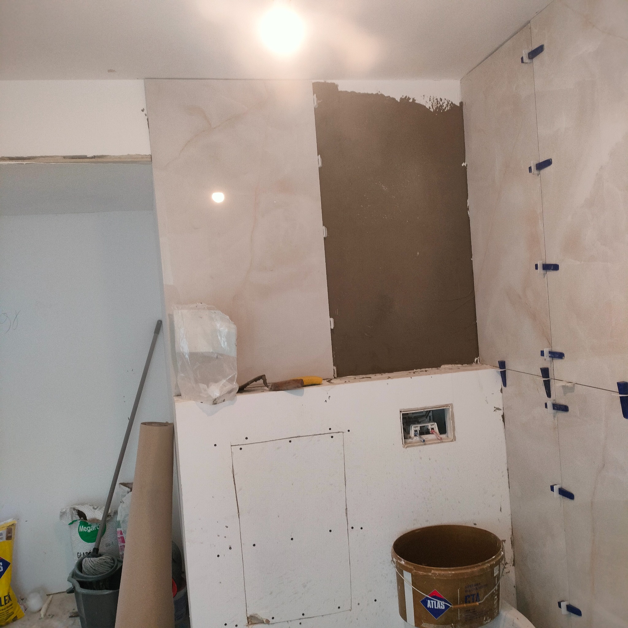 Wnętrze łazienki w trakcie kładzenia płytek. Widoczne narzędzia glazurnicze, wiadro z klejem Atlas oraz płytki z niebieskimi krzyżykami dystansowymi.