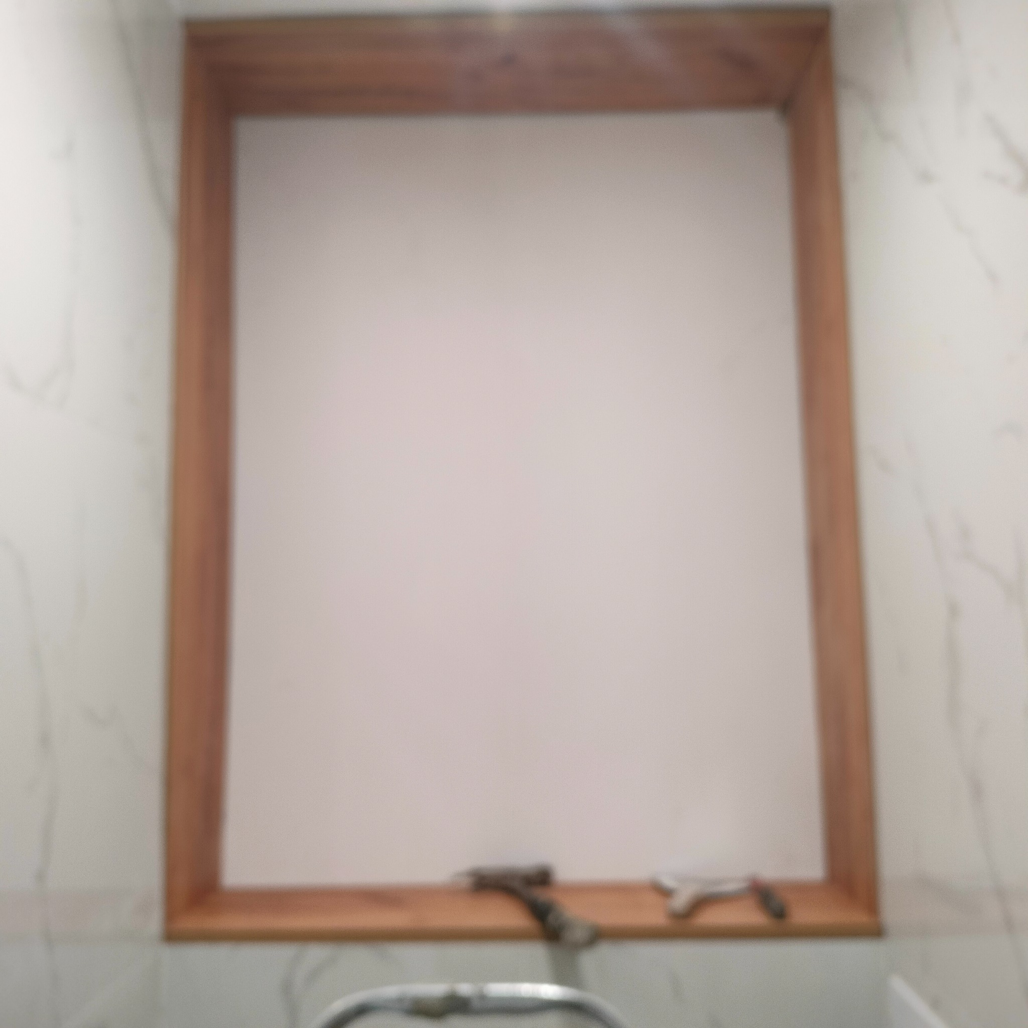 Narożnik ściany z drewnianą ramą i narzędziami glazurniczymi. Płytki imitujące marmur w tle. Fragment baterii umywalkowej widoczny na dole.