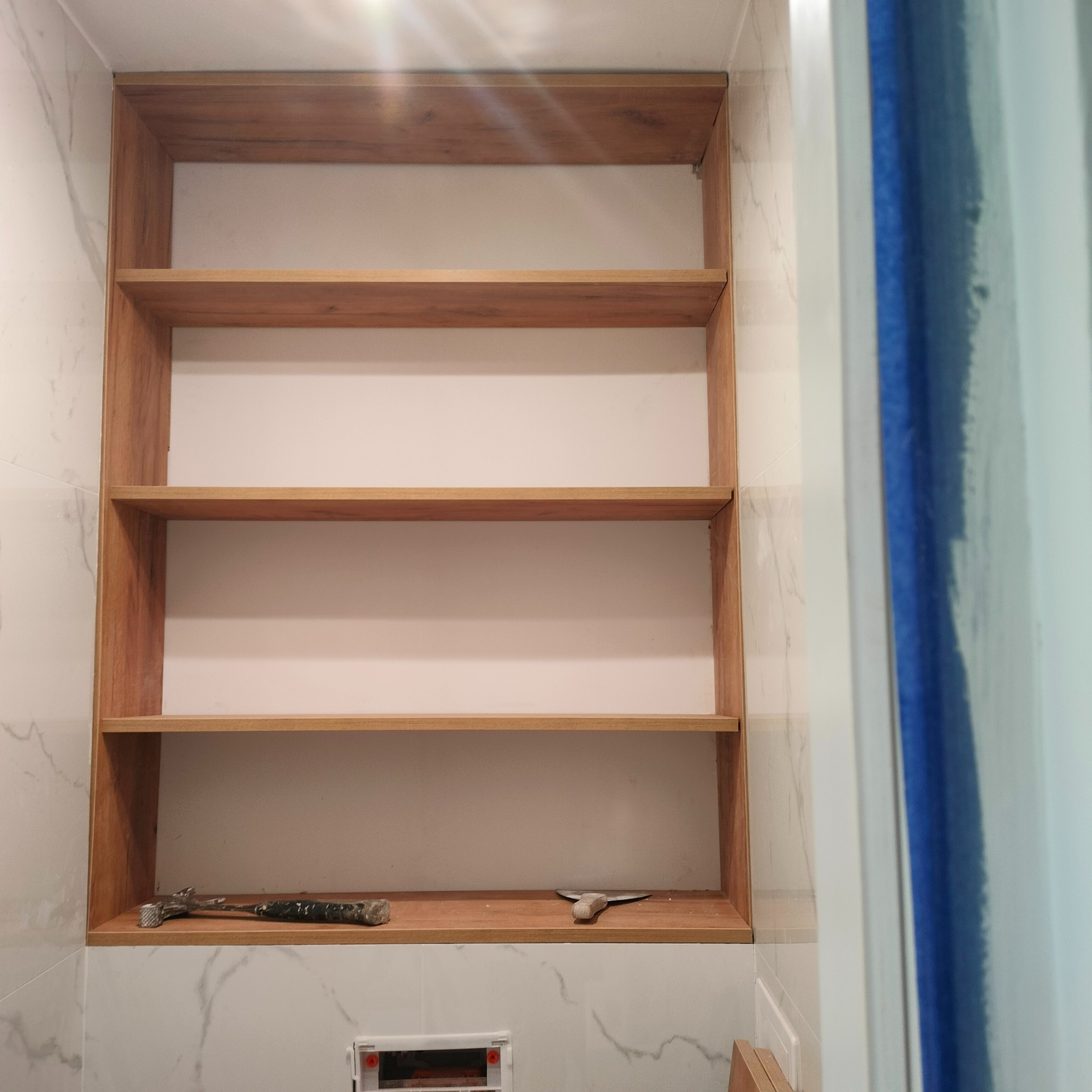 Wnęka z drewnianymi półkami w łazience wykończonej marmurowymi płytkami. Na dolnej półce leżą narzędzia: młotek i szpachelka. Widoczny fragment kabiny prysznicowej.