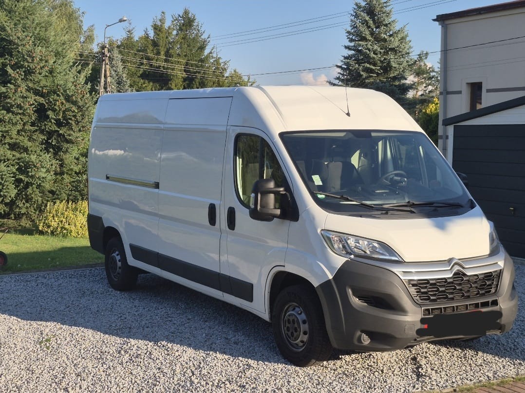 Biały bus dostawczy Citroen Jumper na żwirowym podjeździe, z zielenią w tle i budynkiem. Auto gotowe do transportu, czyste, z anteną na dachu.