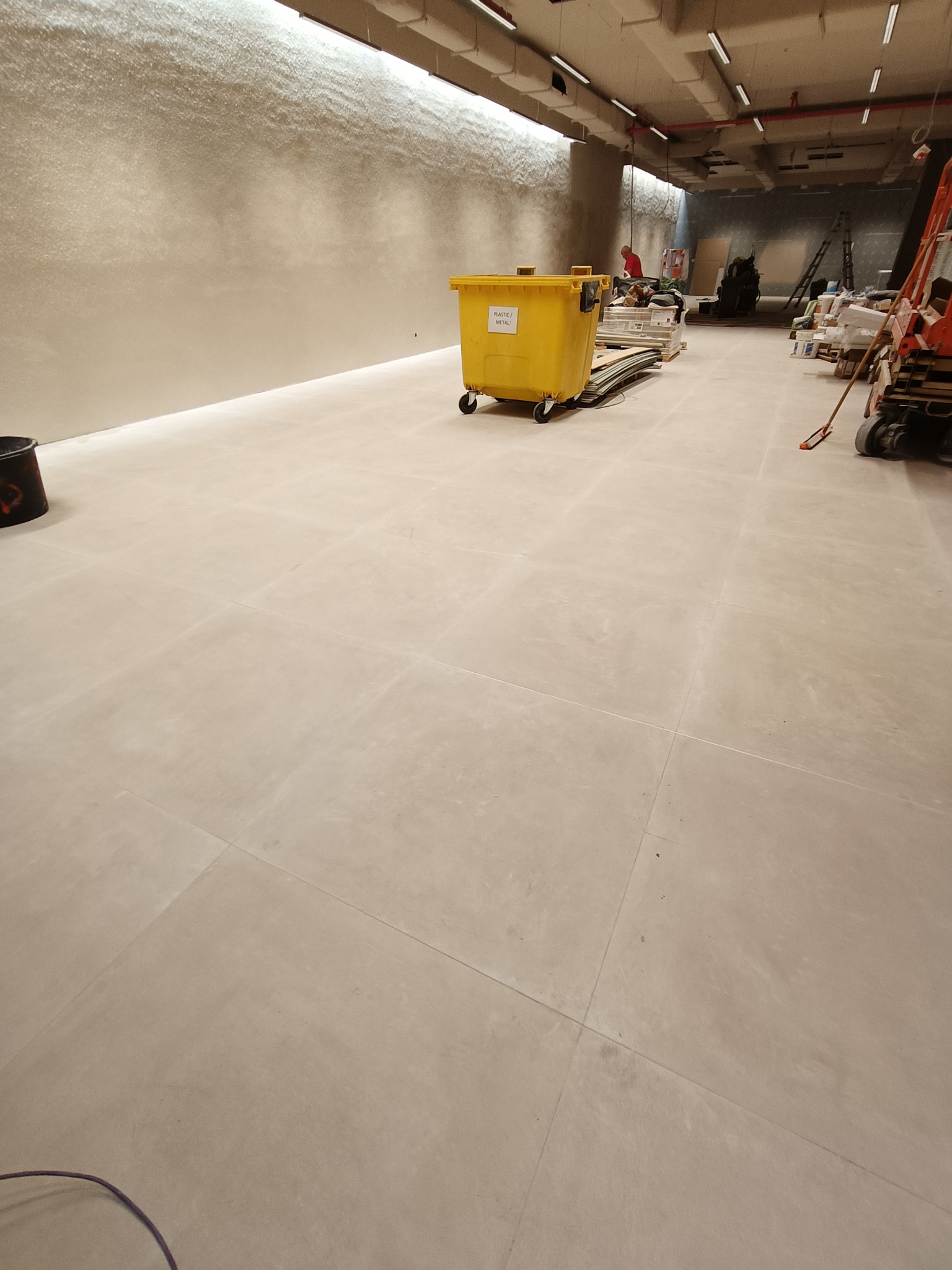 Ułożona jasna, matowa posadzka z płytek wielkoformatowych w trakcie prac wykończeniowych. Widoczny żółty kontener na odpady i narzędzia budowlane w tle.
