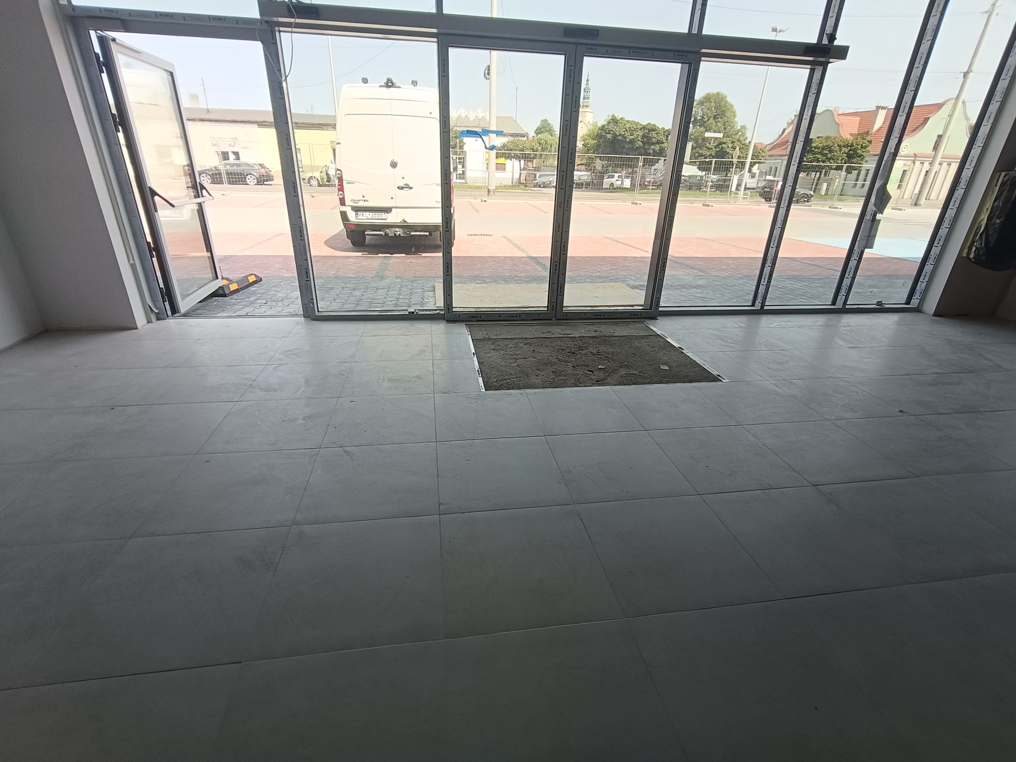 Szare płytki podłogowe w dużym formacie z widocznym miejscem po brakującej płytce, przez przeszklone drzwi widoczny plac z zaparkowanym busem.