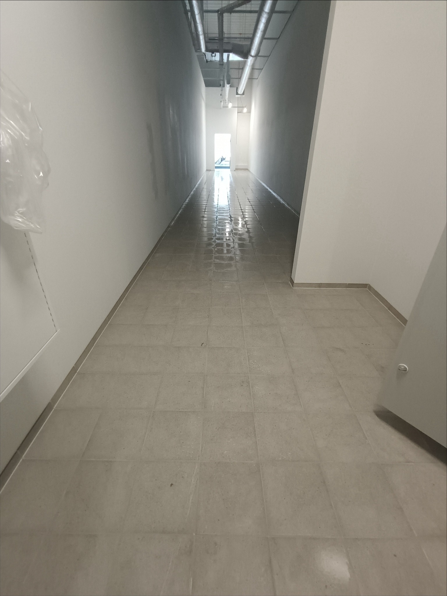 Korytarz z jasnoszarą, świeżo ułożoną terakotą. Widoczne rury pod sufitem i światło dzienne wpadające z końca korytarza. Minimalistyczny design.