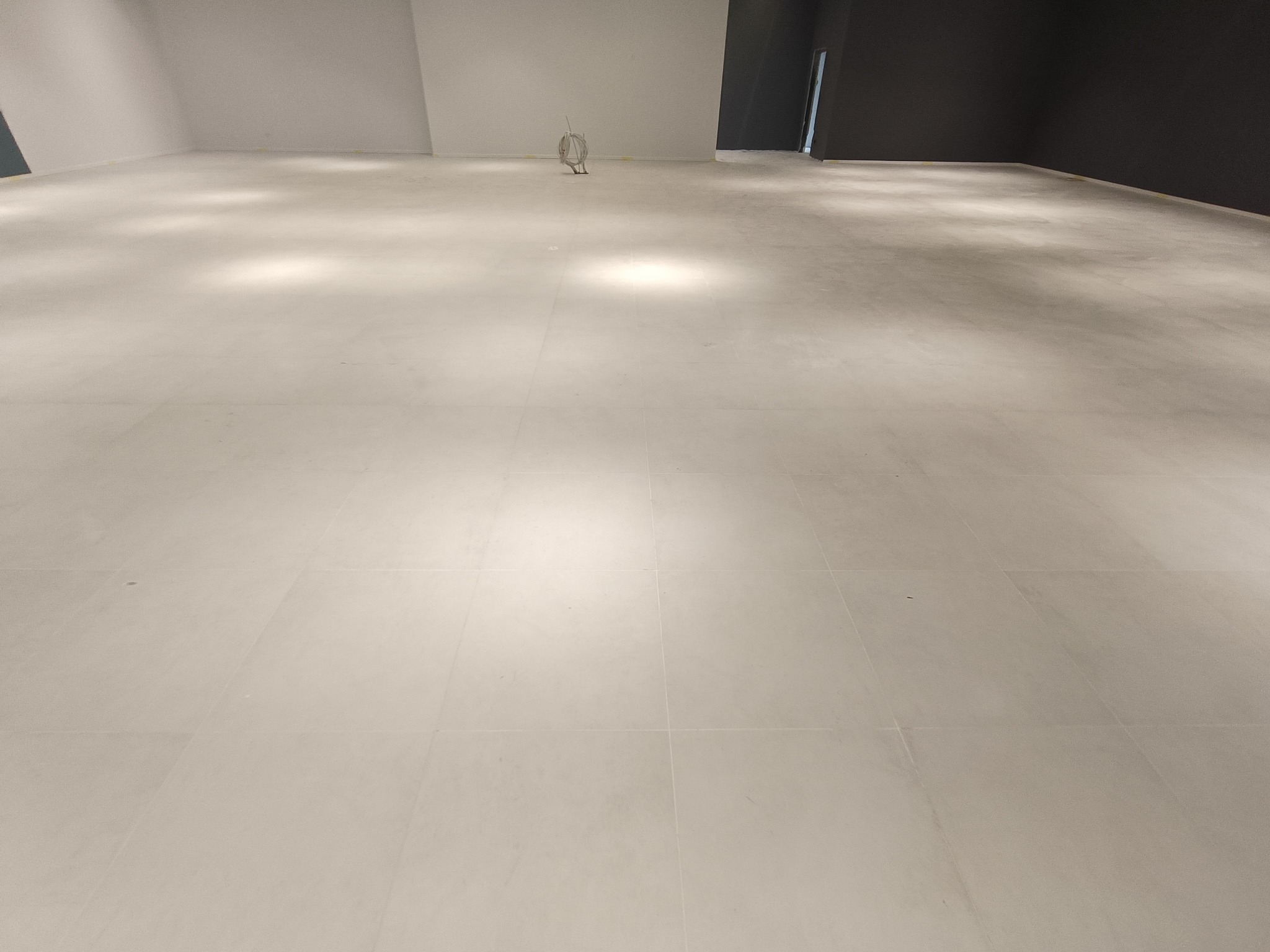 Jasne, duże płytki podłogowe ułożone w przestronnym, minimalistycznym wnętrzu z ciemną ścianą w tle. Widoczne przewody instalacyjne.