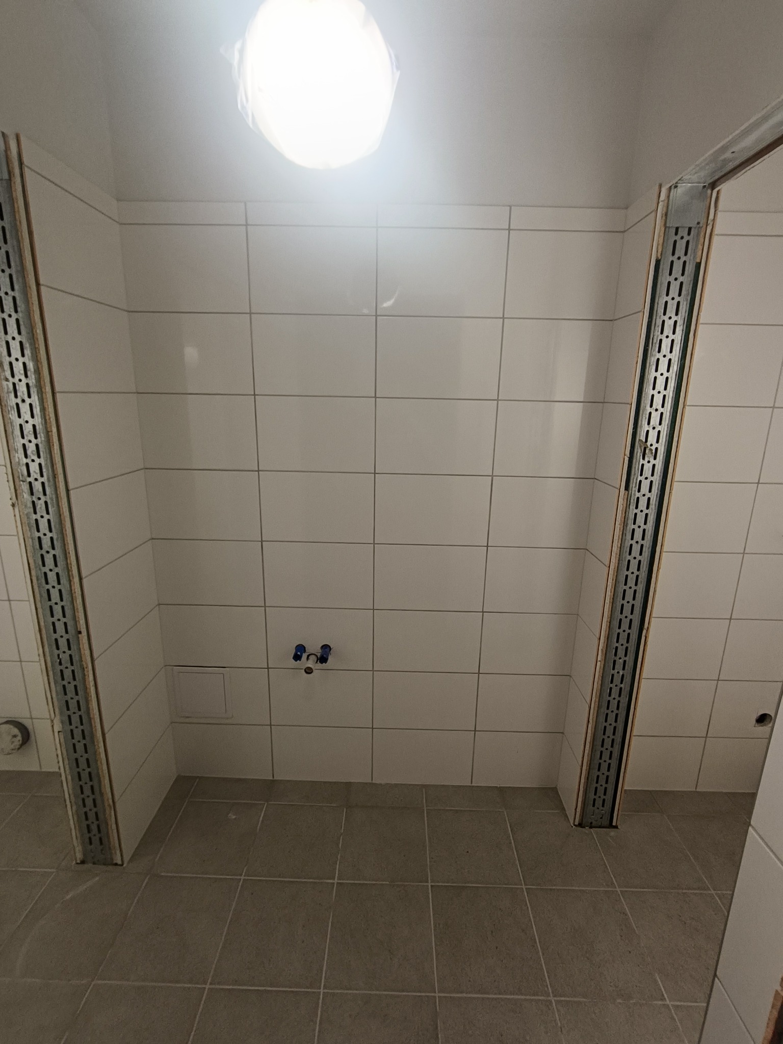 Wnętrze łazienki w trakcie remontu: ułożone jasne płytki na ścianach i podłodze, widoczne metalowe profile w narożnikach, przygotowane przyłącza wodne.