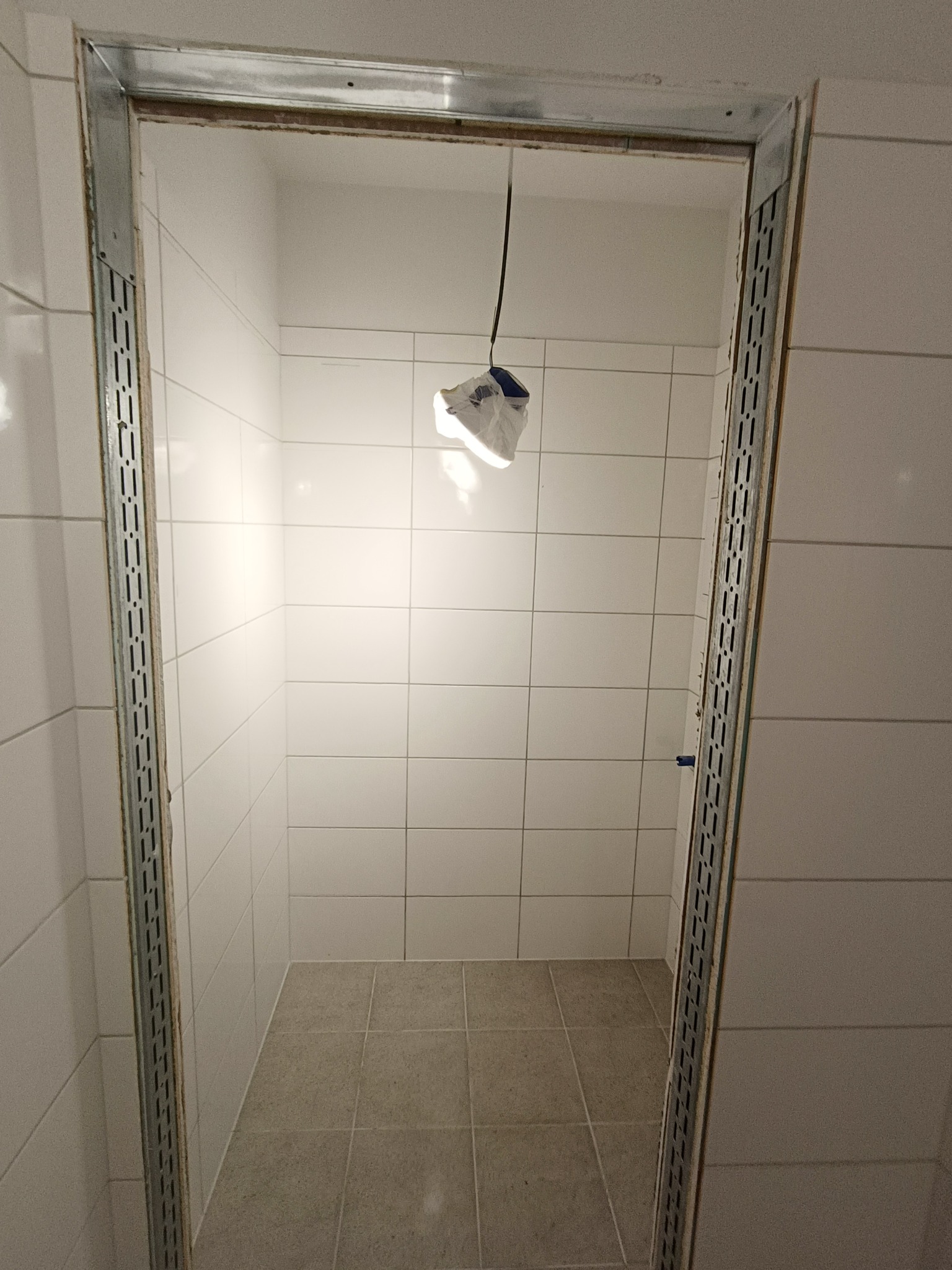 Wykończona kabina prysznicowa z białymi płytkami ściennymi i szarymi podłogowymi. Metalowe profile przy wejściu. Lampa wisząca wewnątrz. Minimalistyczny design.