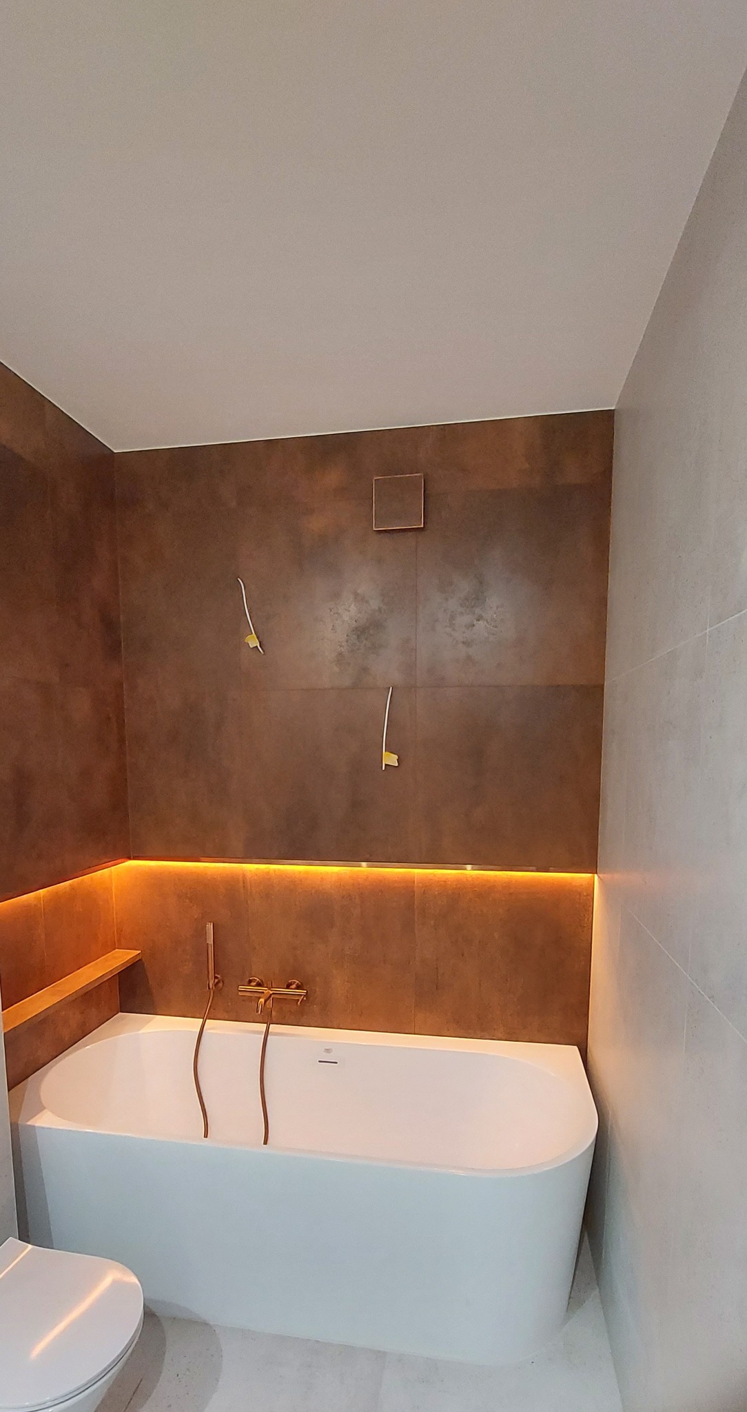 Nowoczesna łazienka z białą wanną, miedzianą armaturą i podświetlanymi półkami. Brązowe płytki imitujące rdzę na ścianie. Widoczne przygotowane punkty pod oświetlenie.
