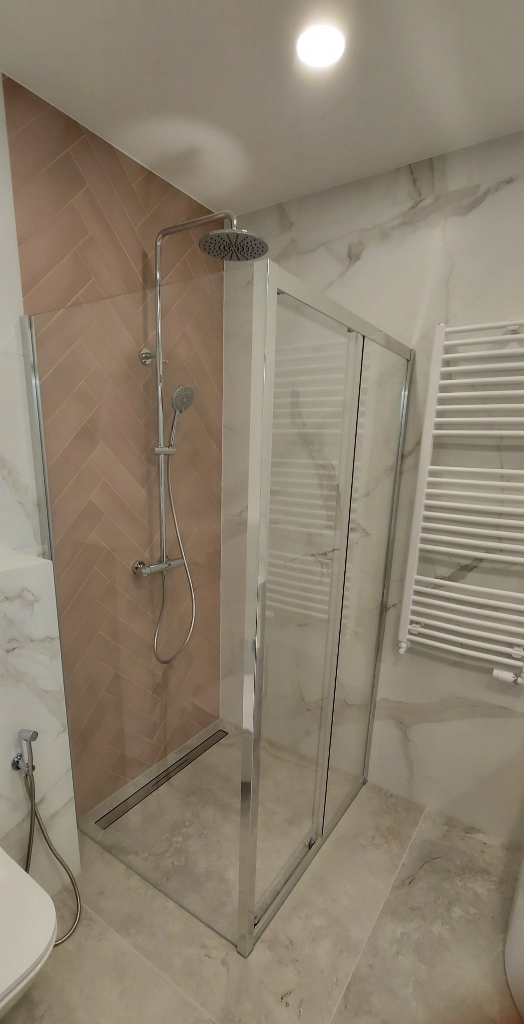 Nowoczesna kabina prysznicowa z deszczownicą i panelami w jodełkę, w łazience wykończonej marmurem. Widoczny grzejnik drabinkowy i fragment toalety.