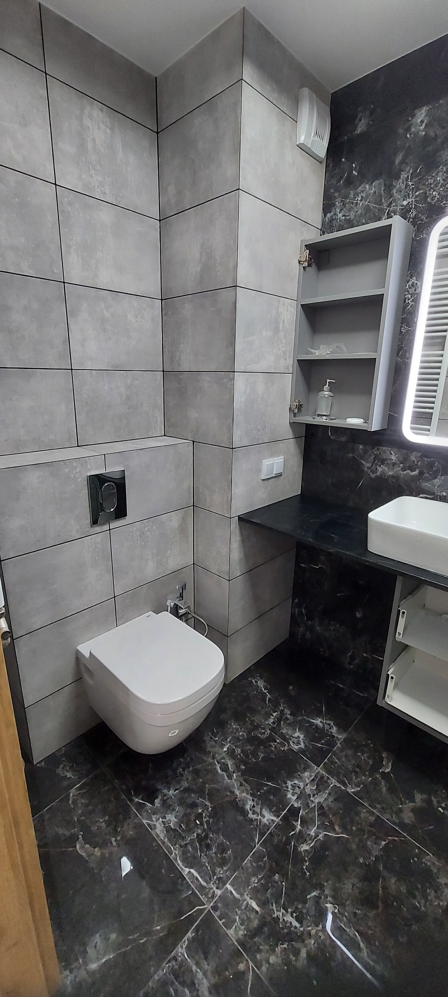 Nowoczesna łazienka z szarymi płytkami ściennymi i czarną marmurową podłogą. Wisząca toaleta, szafka z lustrem i umywalka na blacie.