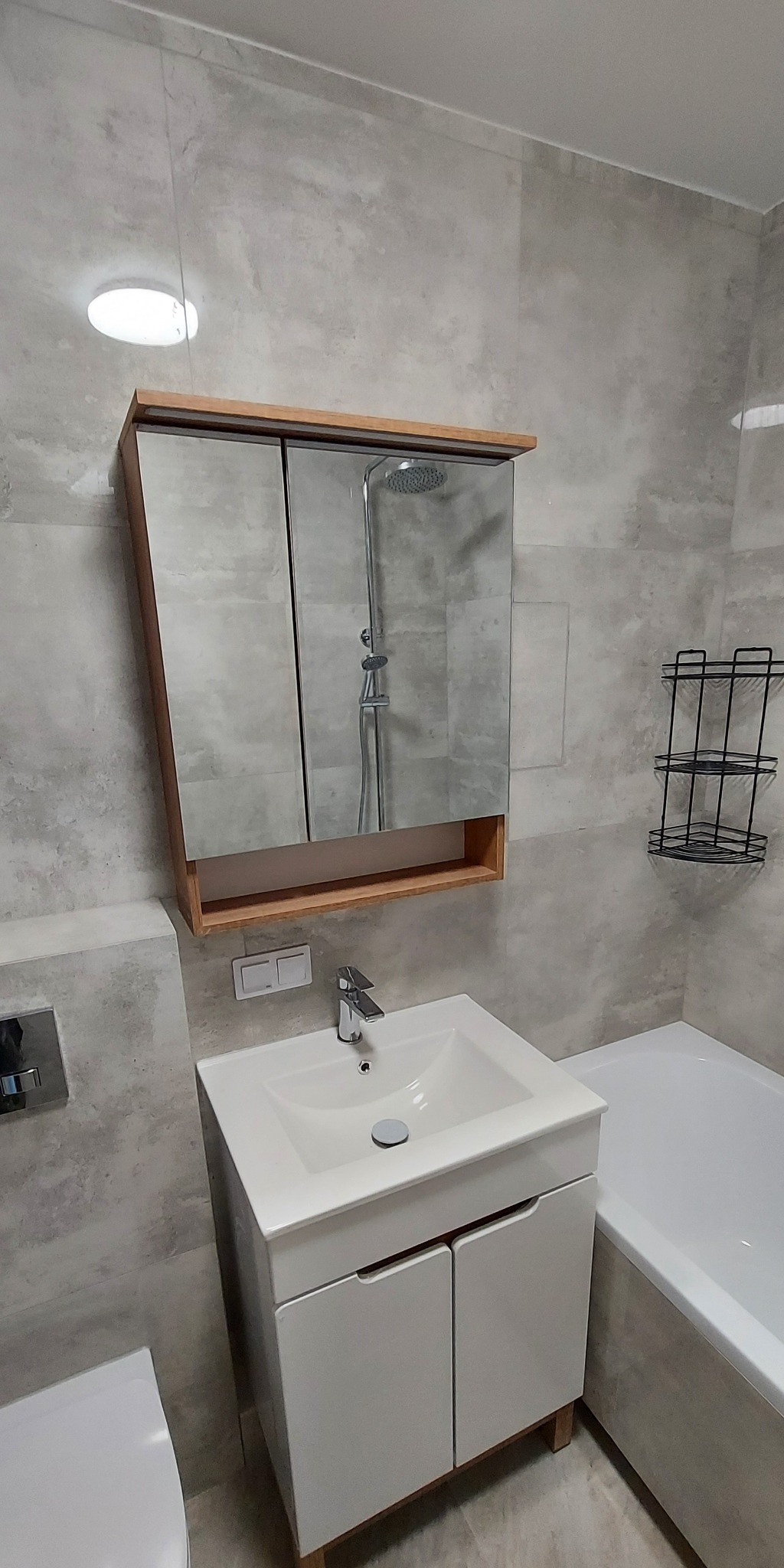 Nowoczesna łazienka z umywalką na szafce, lustrem w drewnianej ramie, szarą glazurą i czarnym metalowym regałem. Widok na kabinę prysznicową w lustrze.