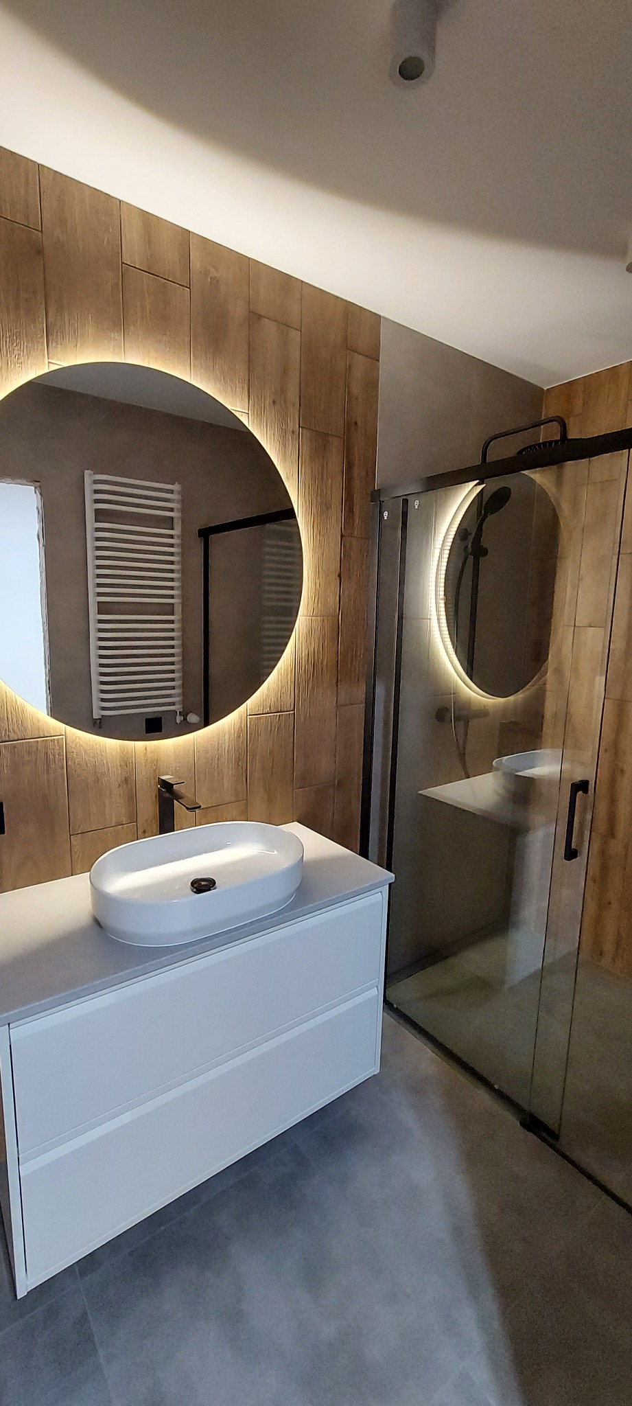 Nowoczesna łazienka z drewnopodobnymi płytkami, okrągłym lustrem z podświetleniem LED, białą szafką z umywalką i kabiną prysznicową z czarnymi detalami.