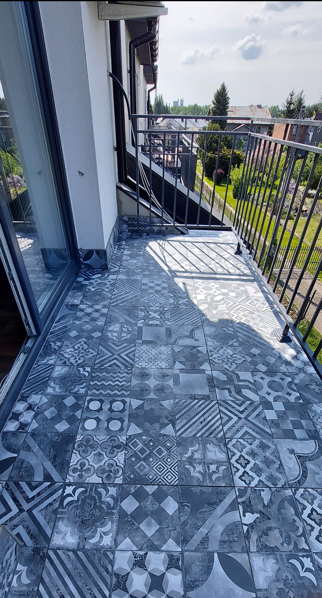 Balkon z szaro-białą, wzorzystą terakotą. Metalowa balustrada, widok na zieleń. Jasne światło słoneczne podkreśla detale płytek i otoczenia.