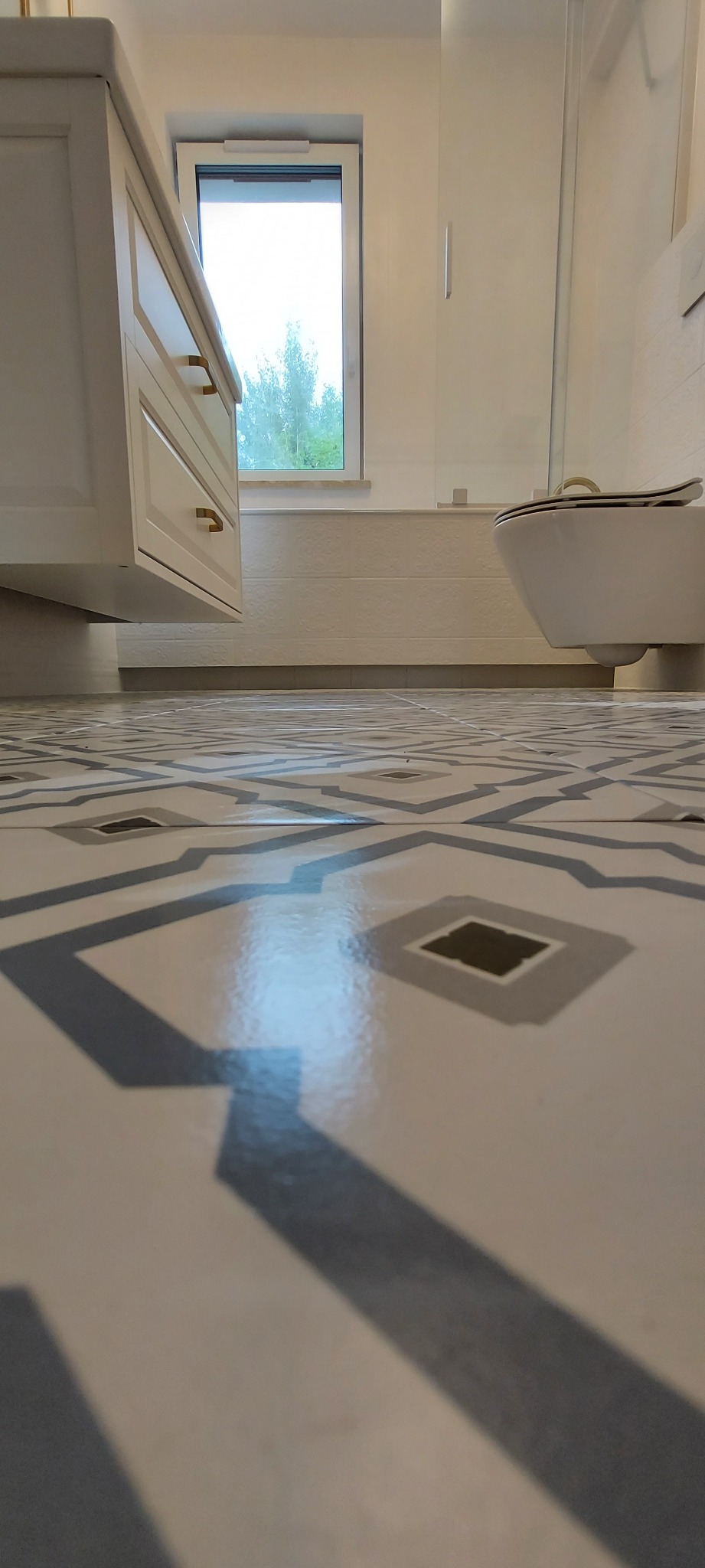 Wnętrze łazienki z wzorzystą, jasną podłogą, białą szafką z złotymi uchwytami, oknem i toaletą. Minimalistyczny design, neutralna kolorystyka.