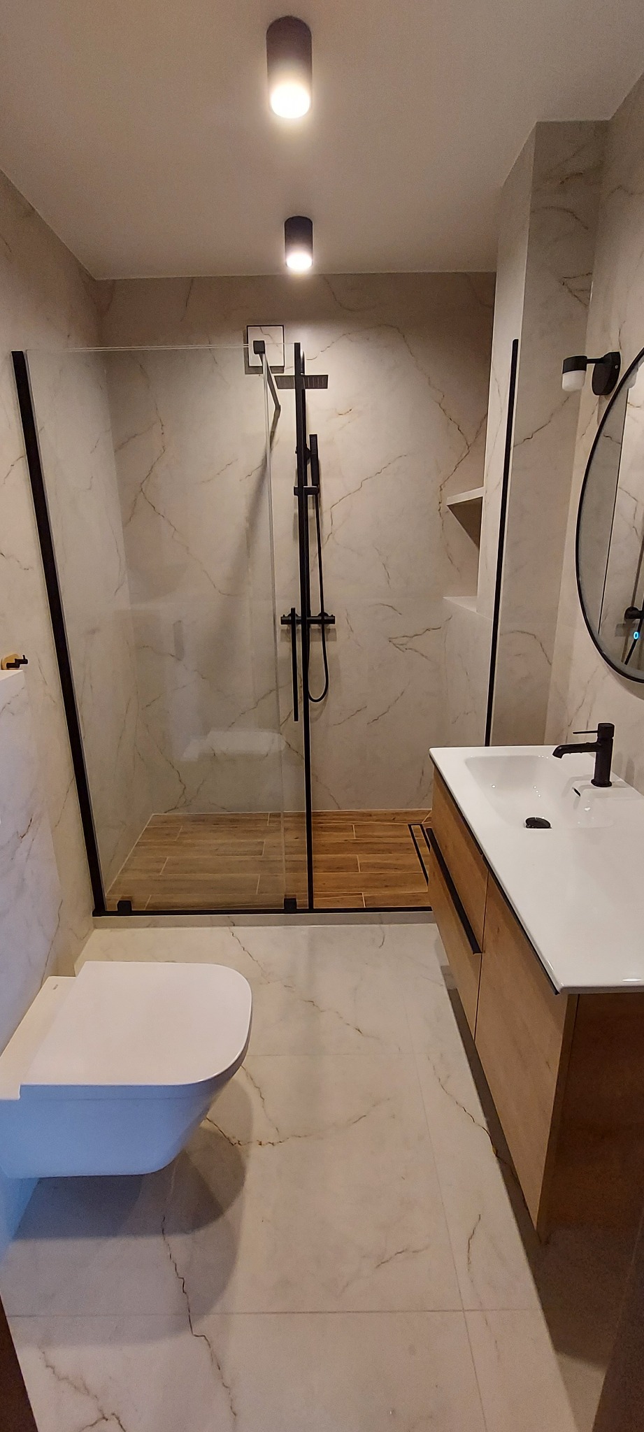 Nowoczesna łazienka z kabiną prysznicową typu walk-in, umywalką na szafce z drewnianym frontem i podwieszaną toaletą. Marmurowe płytki na ścianach i podłodze.