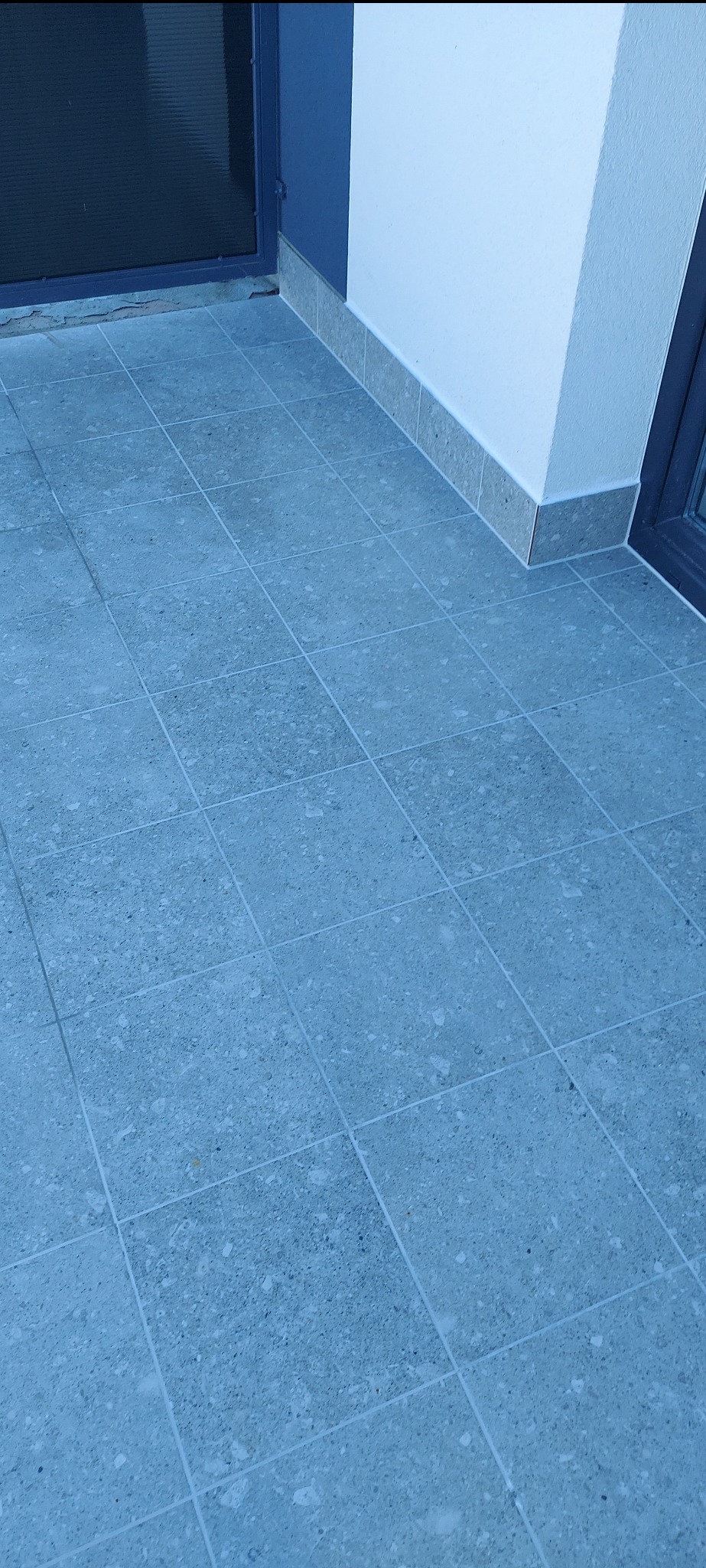 Błękitna, kwadratowa terakota na balkonie, z widocznymi fugami i fragmentem ściany z szarą listwą przypodłogową. Ujęcie z góry, podkreślające wzór płytek.