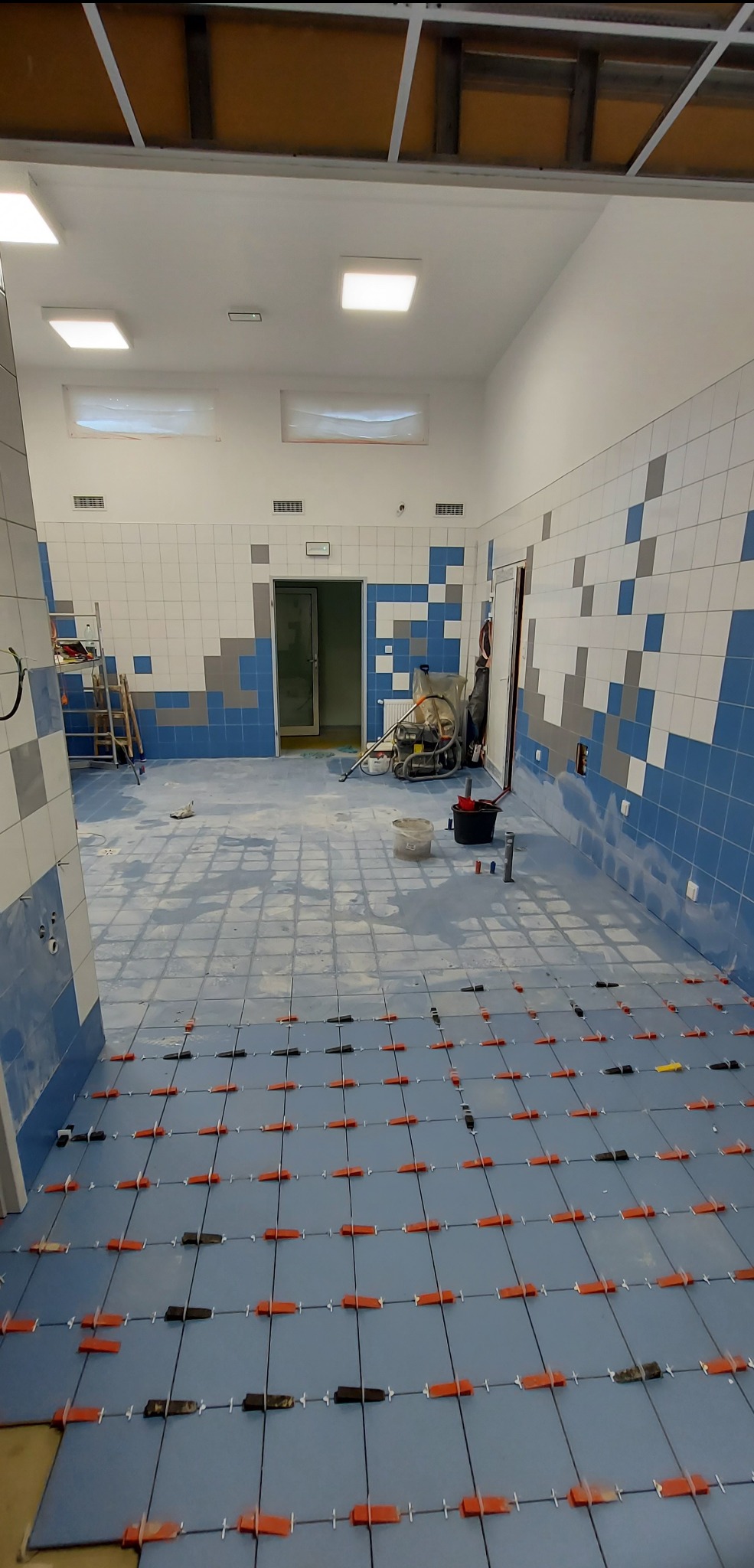 Układanie niebieskich płytek podłogowych z użyciem krzyżyków dystansowych w dużej łazience z częściowo wykończonymi ścianami w biało-niebieską mozaikę.