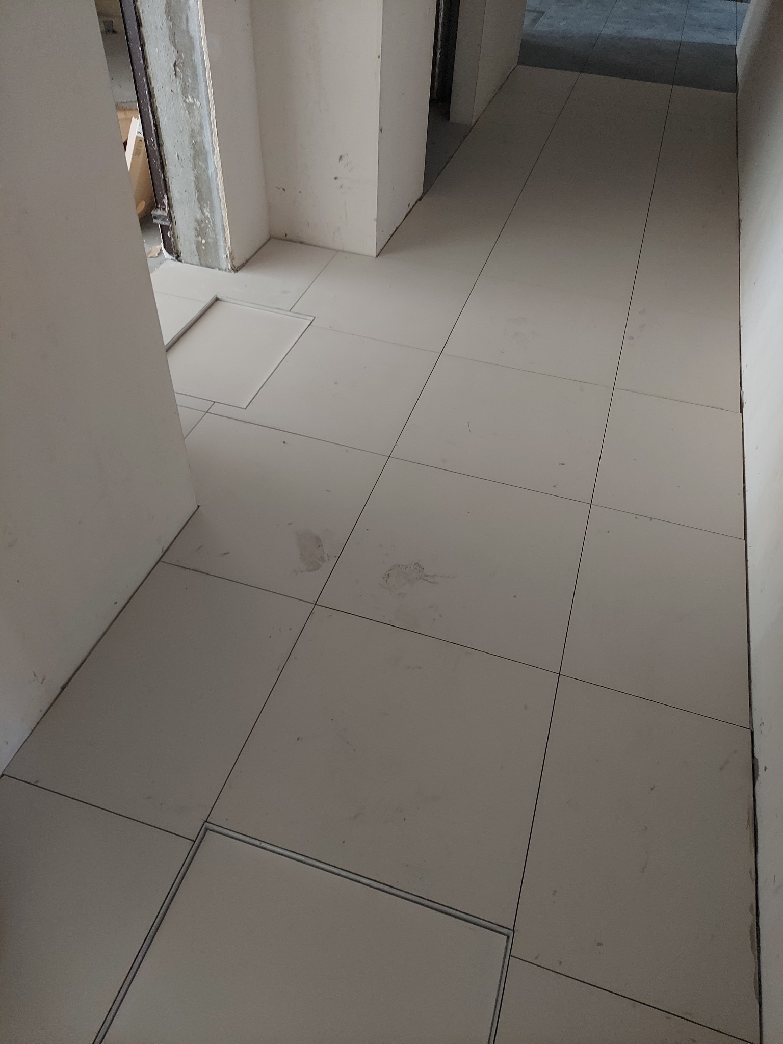 Ułożona, jasna podłoga z płytek w trakcie prac wykończeniowych. Widoczne fragmenty ścian i drzwi. Minimalistyczny styl, surowe wykończenie.