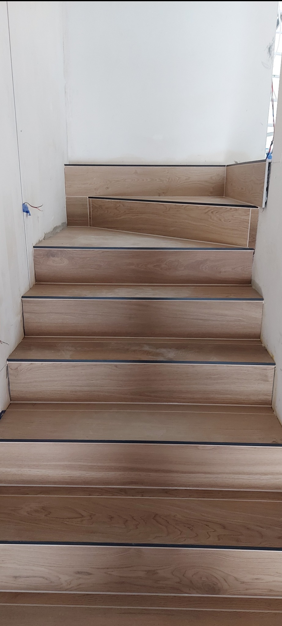 Schody wyłożone płytkami imitującymi drewno, z czarnymi listwami wykończeniowymi. Widoczne fragmenty białych ścian. Ujęcie z dołu, podkreślające perspektywę.