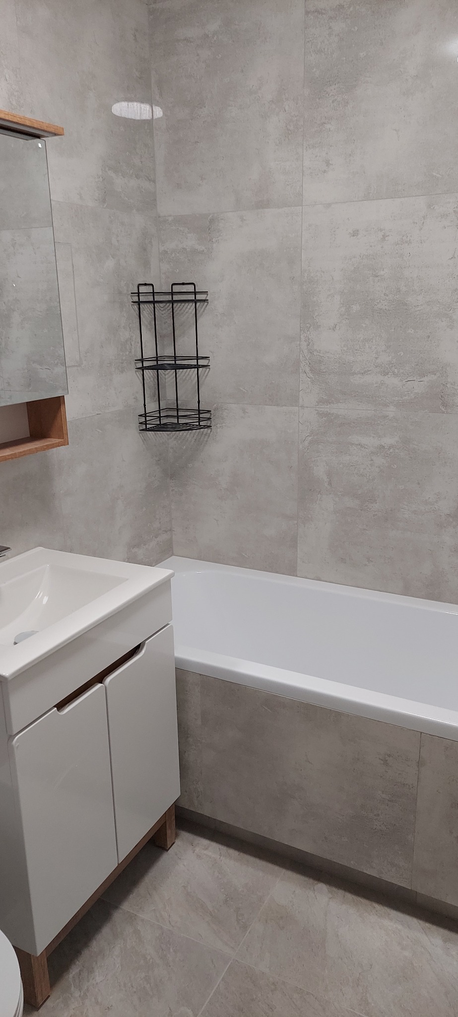 Jasna łazienka po remoncie: biała umywalka z szafką, wanna obudowana płytkami w odcieniach szarości, czarna półka narożna na ścianie z płytek.