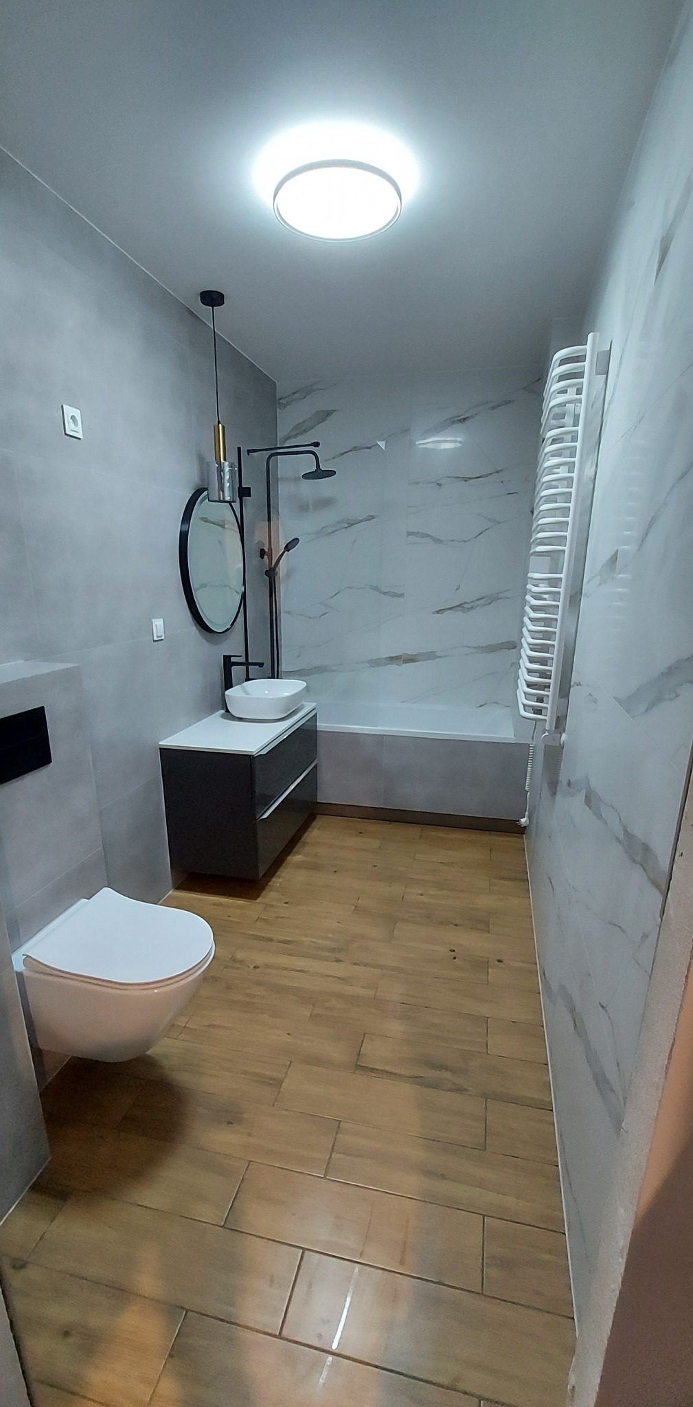 Wykończona łazienka w Krakowie z drewnopodobną podłogą, szarą armaturą i marmurowymi płytkami. Nowoczesny design z okrągłym lustrem i grzejnikiem drabinkowym.