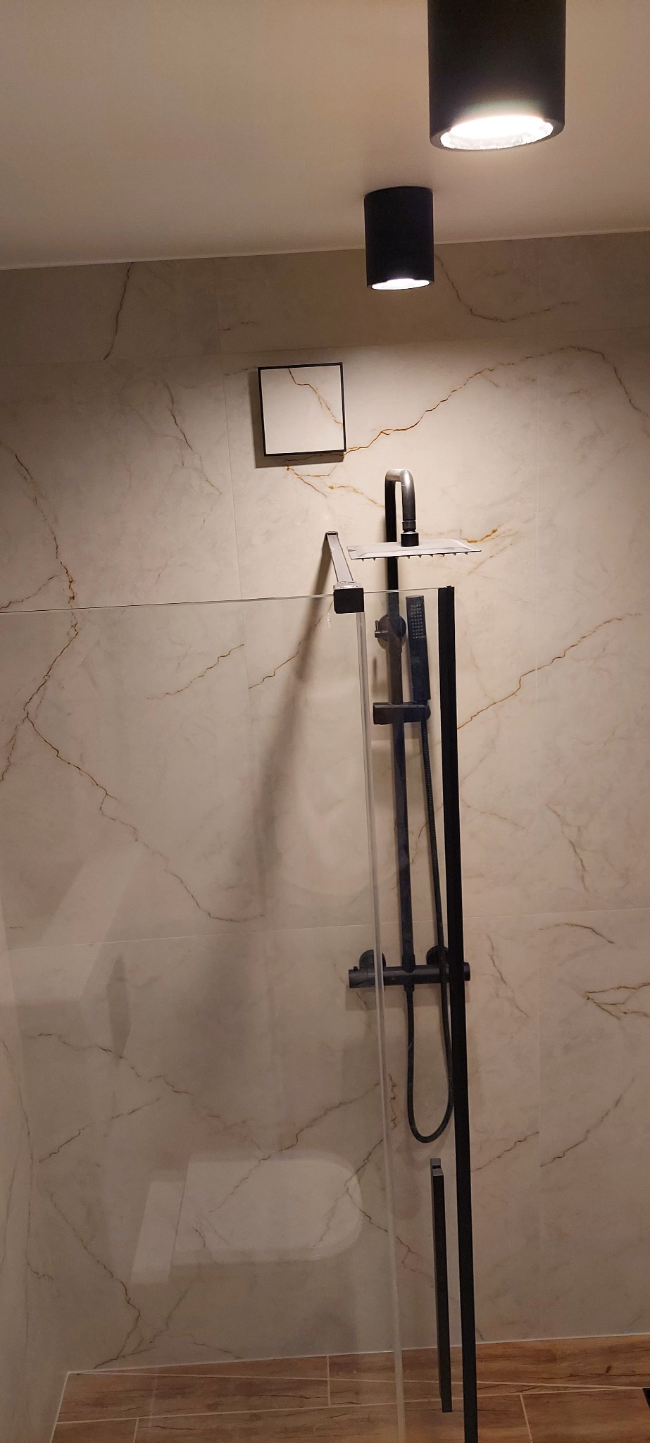 Nowoczesna kabina prysznicowa z czarną armaturą i deszczownicą, marmurowe płytki na ścianach, minimalistyczne oświetlenie sufitowe. Wykończenie łazienki w eleganckim stylu.