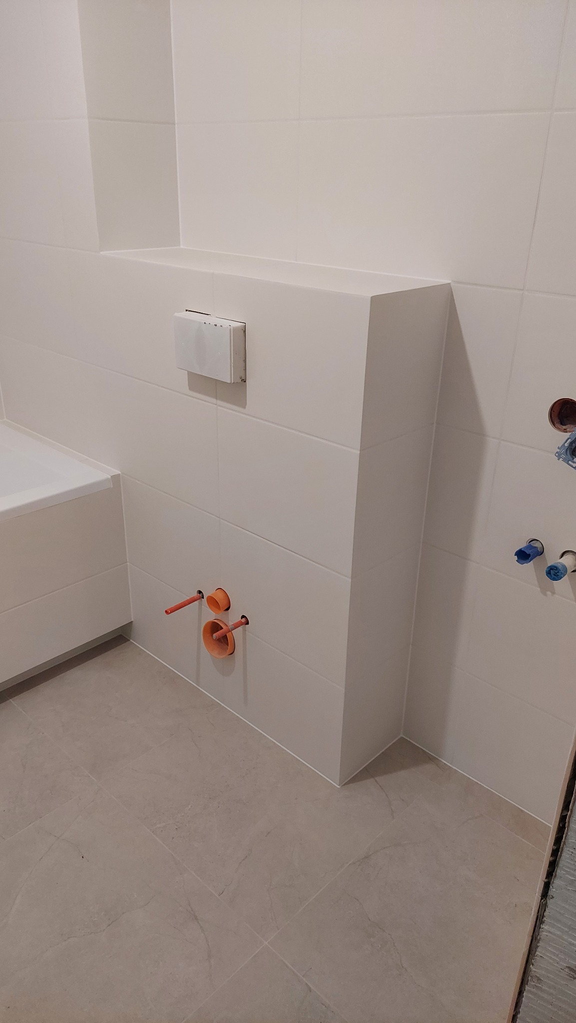 Wykończona łazienka z zabudową geberitu i białymi płytkami. Widoczne wystające rury instalacji wodnej i kanalizacyjnej. Na podłodze szare płytki imitujące kamień.