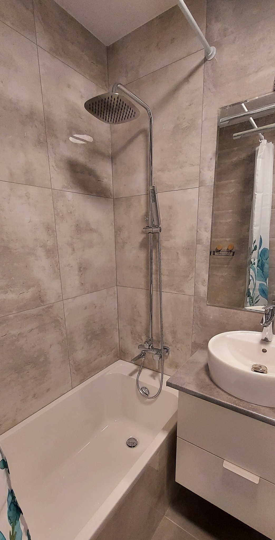 Jasna łazienka po remoncie: wanna z deszczownicą, umywalka z szafką i lustro. Płytki w odcieniach szarości. Minimalistyczny design.