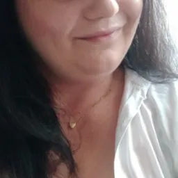 Portret kobiety z ciemnymi włosami, uśmiechającej się do kamery. Widoczny złoty łańcuszek z serduszkiem. Ubrana w białą koszulę i czarny top.