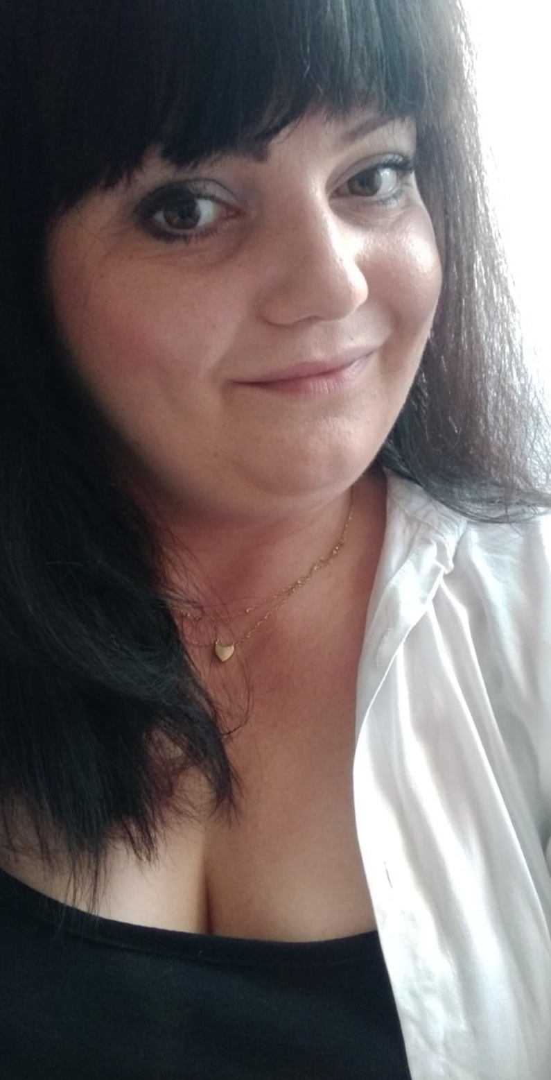 Portret kobiety z ciemnymi włosami, uśmiechającej się do kamery. Widoczny złoty łańcuszek z serduszkiem. Ubrana w białą koszulę i czarny top.